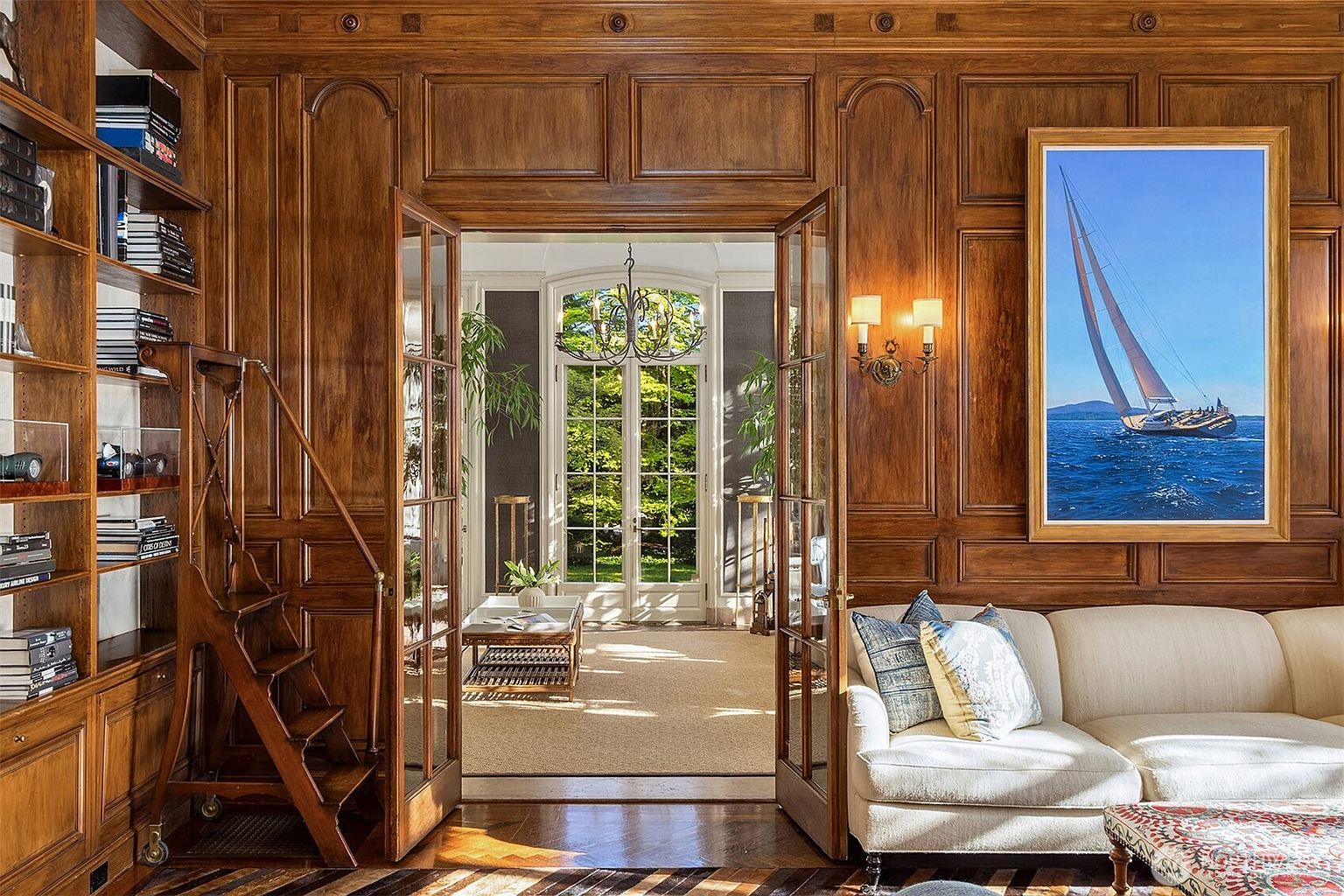 This sophisticated study features rich, floor-to-ceiling wood paneling and built-in bookshelves complete with a rolling library ladder. A plush, cream-colored sofa sits beneath a large, vibrant painting of a sailboat, while glass-paned double doors open into a bright, sun-drenched conservatory beyond. The space exudes a classic, scholarly elegance, perfectly blending traditional craftsmanship with a serene, airy transition to the outdoors.