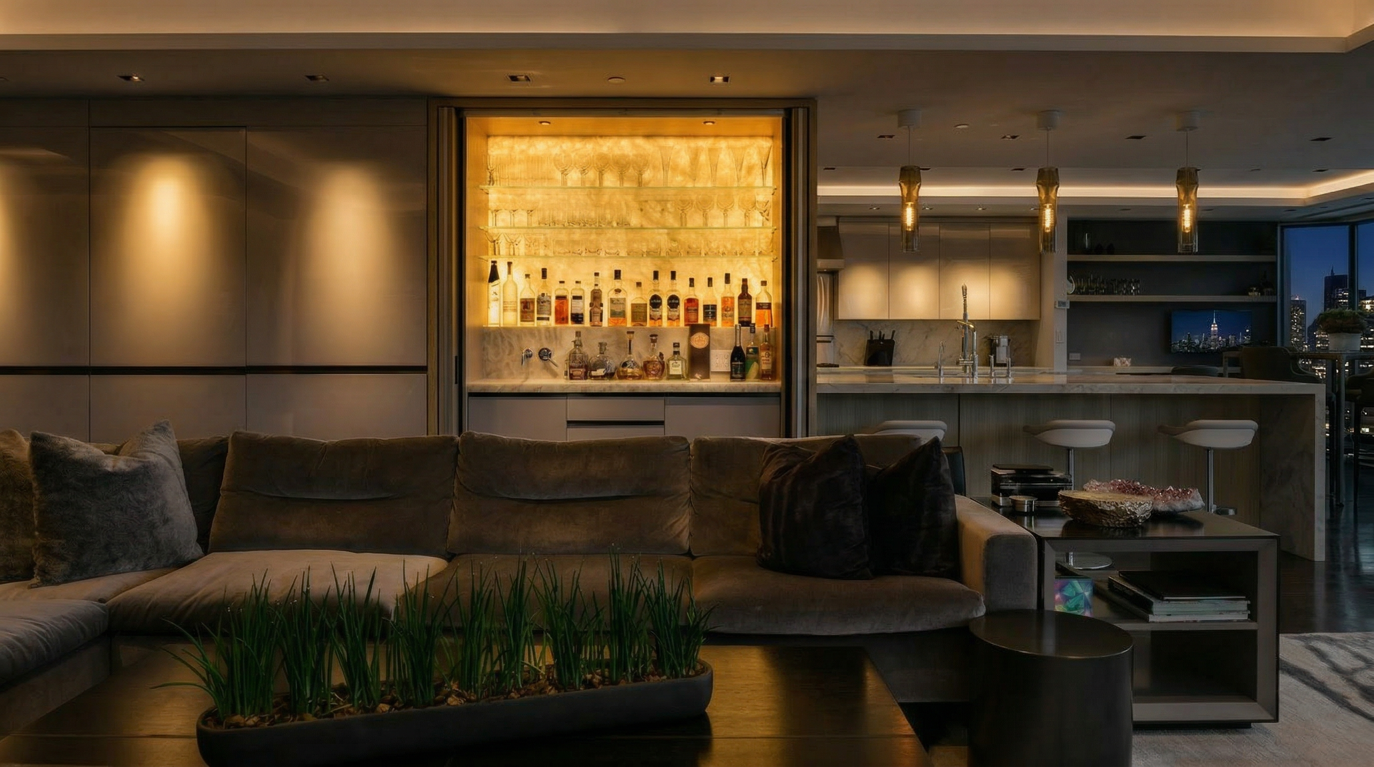 This sophisticated and modern living area features a plush, dark neutral sectional sofa as its centerpiece, facing a built-in illuminated bar display. The space flows seamlessly into a contemporary kitchen and dining area, highlighted by elegant pendant lighting and warm ambient backlighting. The open-concept layout and refined finishes lend the room a luxurious, high-end atmosphere suitable for both relaxation and entertaining.