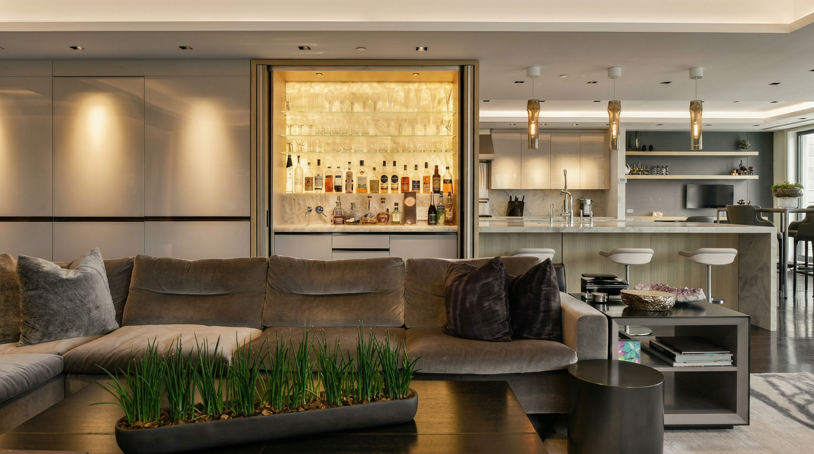 This sophisticated open-concept living space features a plush, velvet-upholstered sectional sofa centered around a dark coffee table adorned with a modern grass planter. A built-in, illuminated bar cabinet serves as a striking focal point, seamlessly transitioning into a sleek, contemporary kitchen with marble countertops and bar seating. The overall aesthetic is one of refined luxury, characterized by neutral tones, ambient recessed lighting, and high-end finishes.