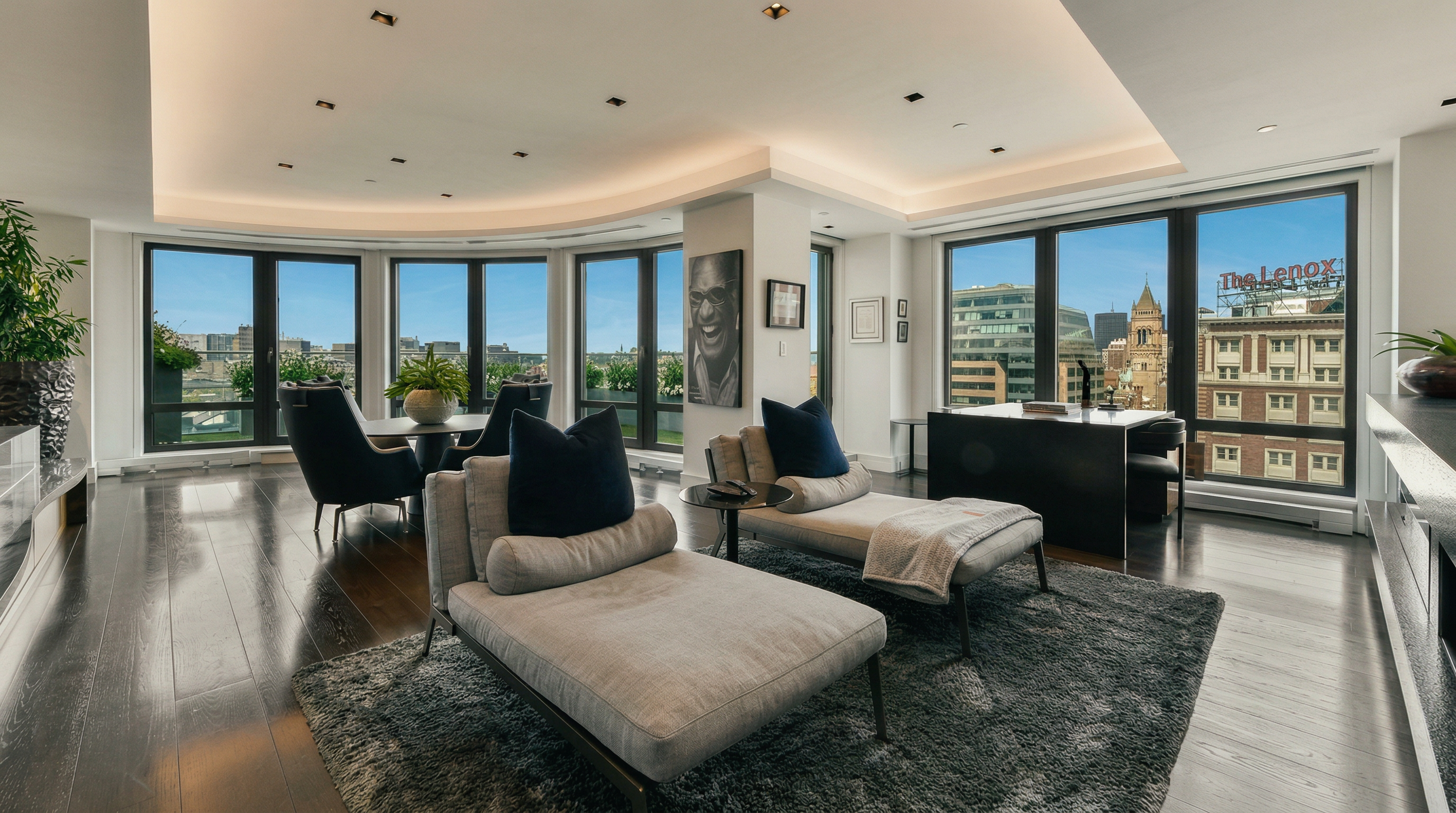 This sophisticated, open-concept living space features a contemporary design with dark hardwood floors and a large, plush area rug anchoring two modern chaise lounges. The room is bathed in natural light from floor-to-ceiling windows that offer expansive city views, including a prominent view of The Lenox building. A sleek desk area and a dining nook are seamlessly integrated into the layout, creating a versatile and luxurious urban living environment.