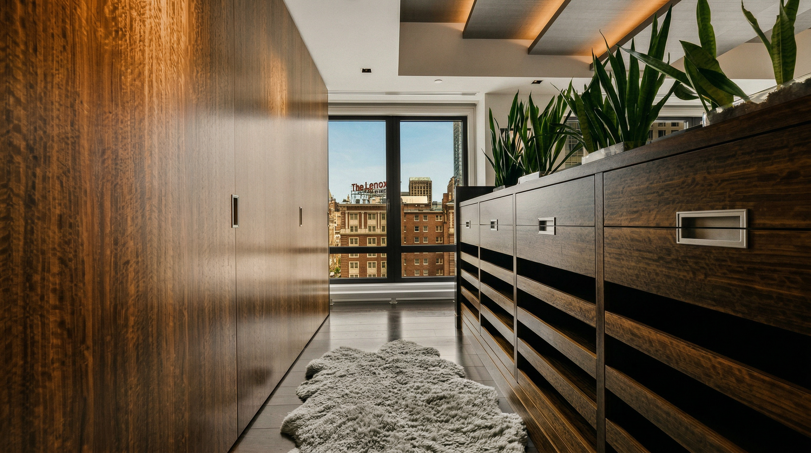 This sophisticated walk-in closet features rich, dark wood cabinetry that spans the length of the room, providing ample storage and a luxurious aesthetic. A plush, white faux-fur rug centers the space, while a row of potted snake plants adds a touch of organic greenery atop the shelving. The perspective looks down the narrow corridor toward a large window that frames a vibrant urban cityscape, blending high-end interior design with a dramatic metropolitan backdrop.