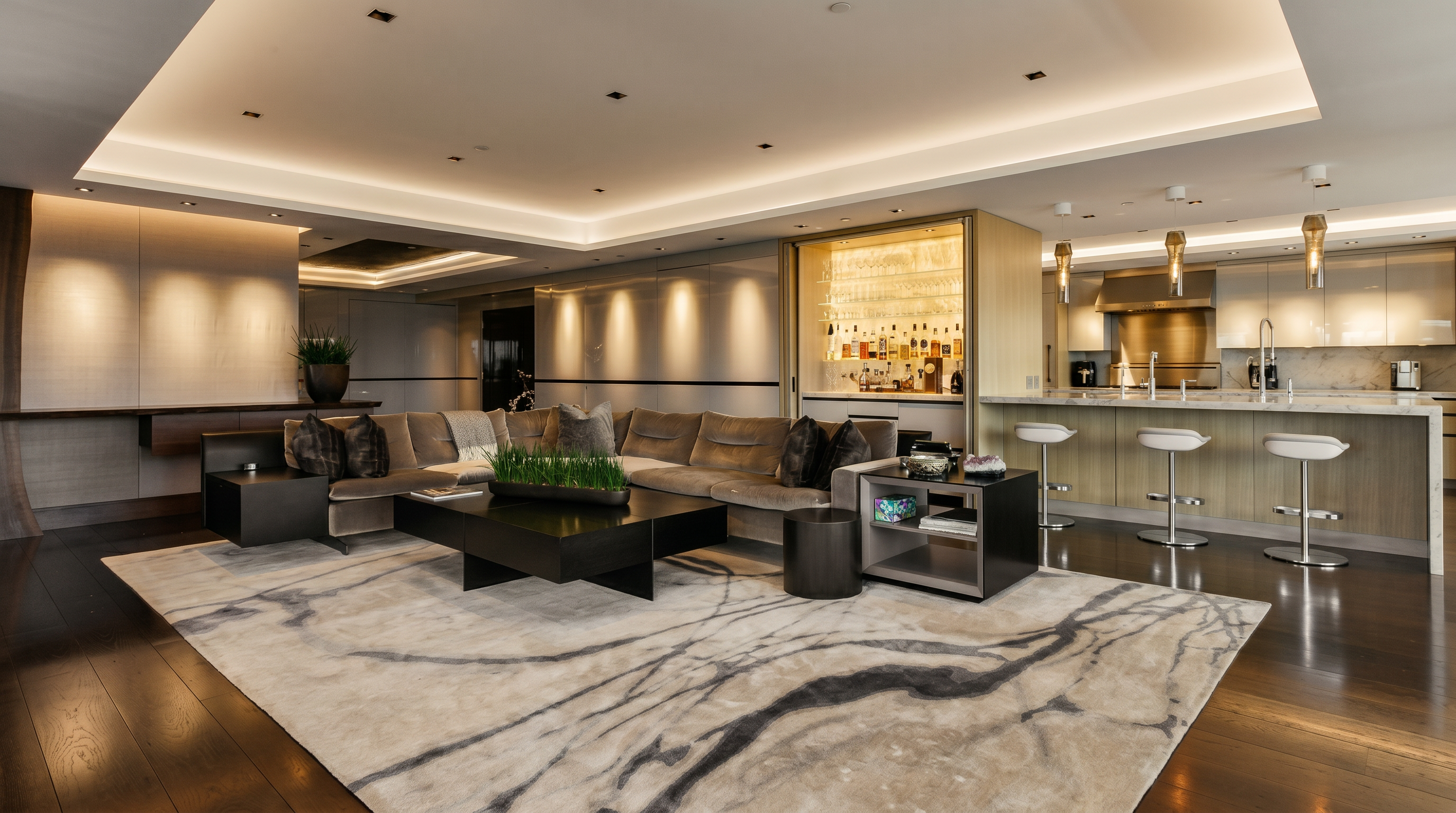 This sophisticated open-concept living area features a plush, L-shaped sectional sofa centered around a bold, black geometric coffee table atop a striking abstract area rug. The space seamlessly integrates a modern kitchen with a breakfast bar and a built-in illuminated bar cabinet, all highlighted by recessed ceiling lighting and warm ambient wall sconces. The dark hardwood flooring and neutral color palette create a luxurious, contemporary atmosphere perfect for high-end entertaining.