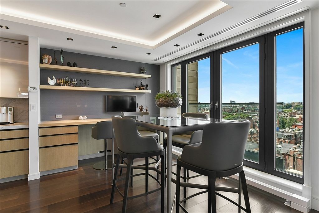 This modern dining area features a sleek glass-topped table surrounded by four contemporary grey upholstered bar stools, set against a backdrop of dark wood flooring and a sophisticated grey accent wall with built-in shelving. Large floor-to-ceiling windows provide an expansive, bright view of the city skyline, while recessed lighting and a minimalist design aesthetic create an elegant, high-end atmosphere. The space seamlessly integrates a functional workspace with a wall-mounted television, making it a versatile area for both dining and professional tasks.
