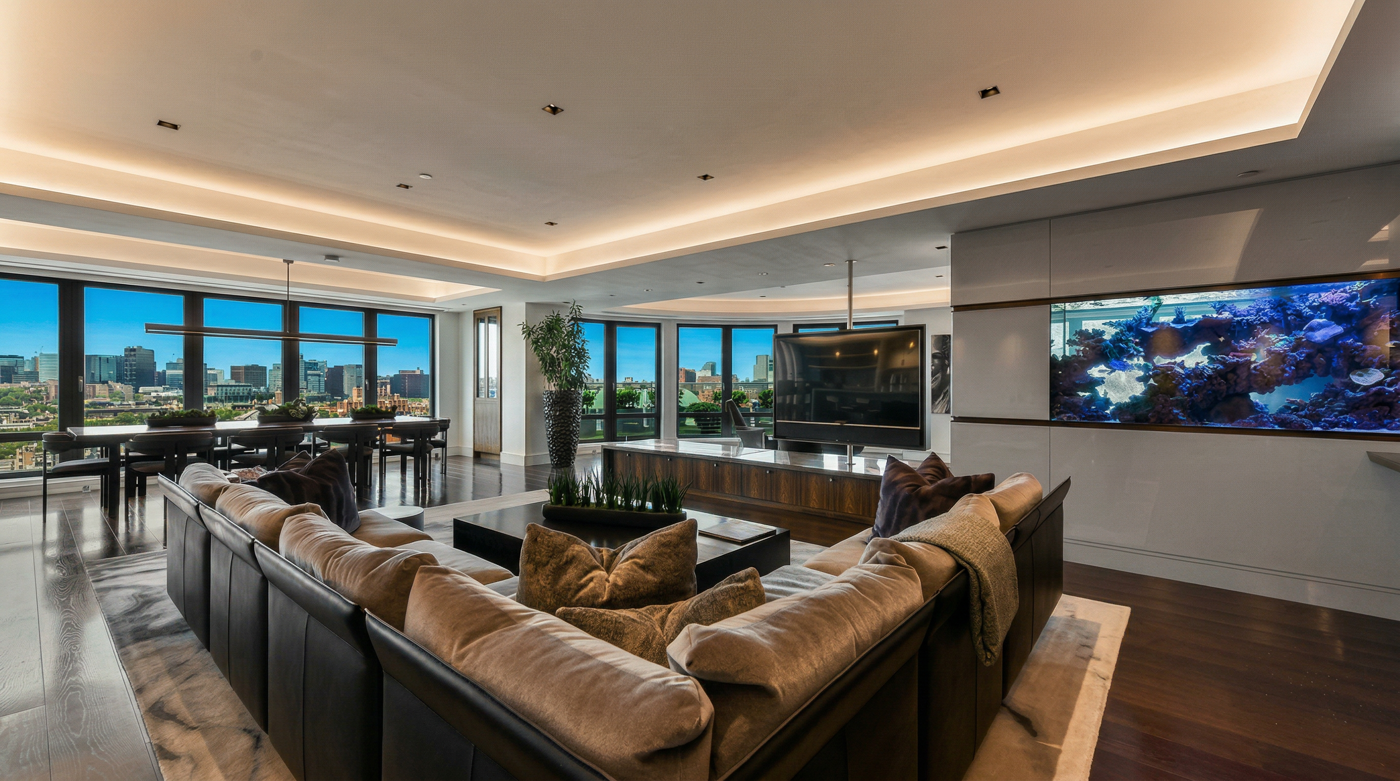 This expansive, modern living room features a large, plush sectional sofa arranged around a central coffee table, facing a sleek television mounted on a custom wooden console. A striking, illuminated built-in aquarium serves as a focal point on the wall, while floor-to-ceiling windows provide panoramic city views and abundant natural light. The open-concept layout seamlessly connects the living area to a formal dining space, creating an atmosphere of sophisticated luxury and comfort.