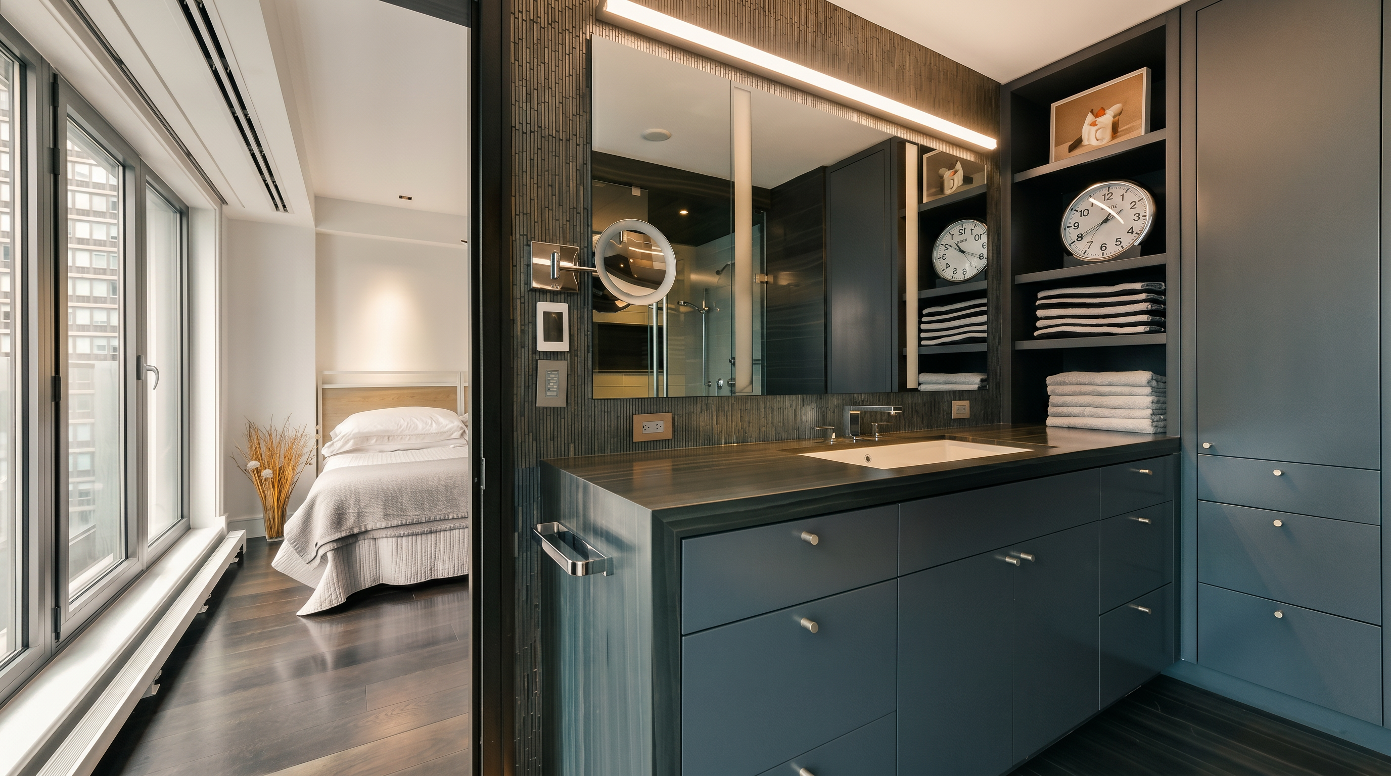 This sophisticated primary bathroom features a sleek, dark grey vanity with a clean-lined countertop and integrated sink, complemented by open shelving stocked with neatly folded towels. A large, illuminated mirror reflects the adjacent bedroom, creating a seamless flow between the two spaces. The modern, minimalist aesthetic is enhanced by dark wood-toned flooring and textured wall accents, offering a luxurious and contemporary feel.