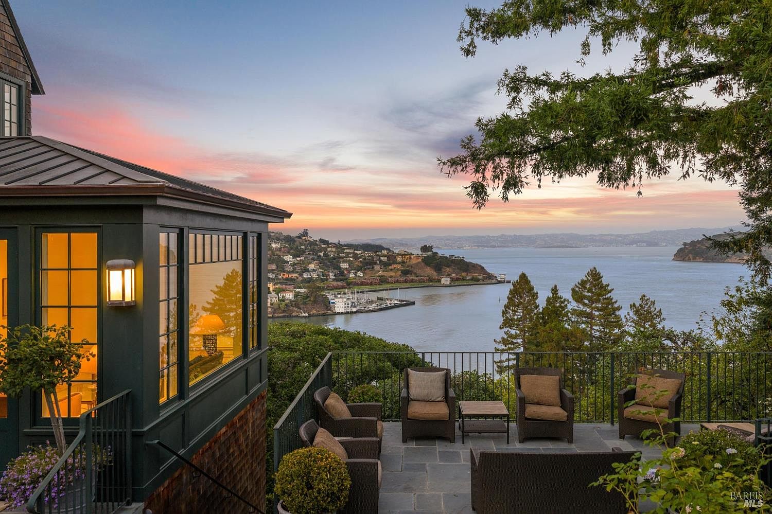 This image showcases a luxurious patio or deck area with stunning waterfront views. The space is furnished with comfortable outdoor seating, creating an inviting atmosphere for relaxation and entertainment. The architecture of the house is visible on the left, and the overall impression is one of upscale living with a focus on enjoying the natural surroundings.