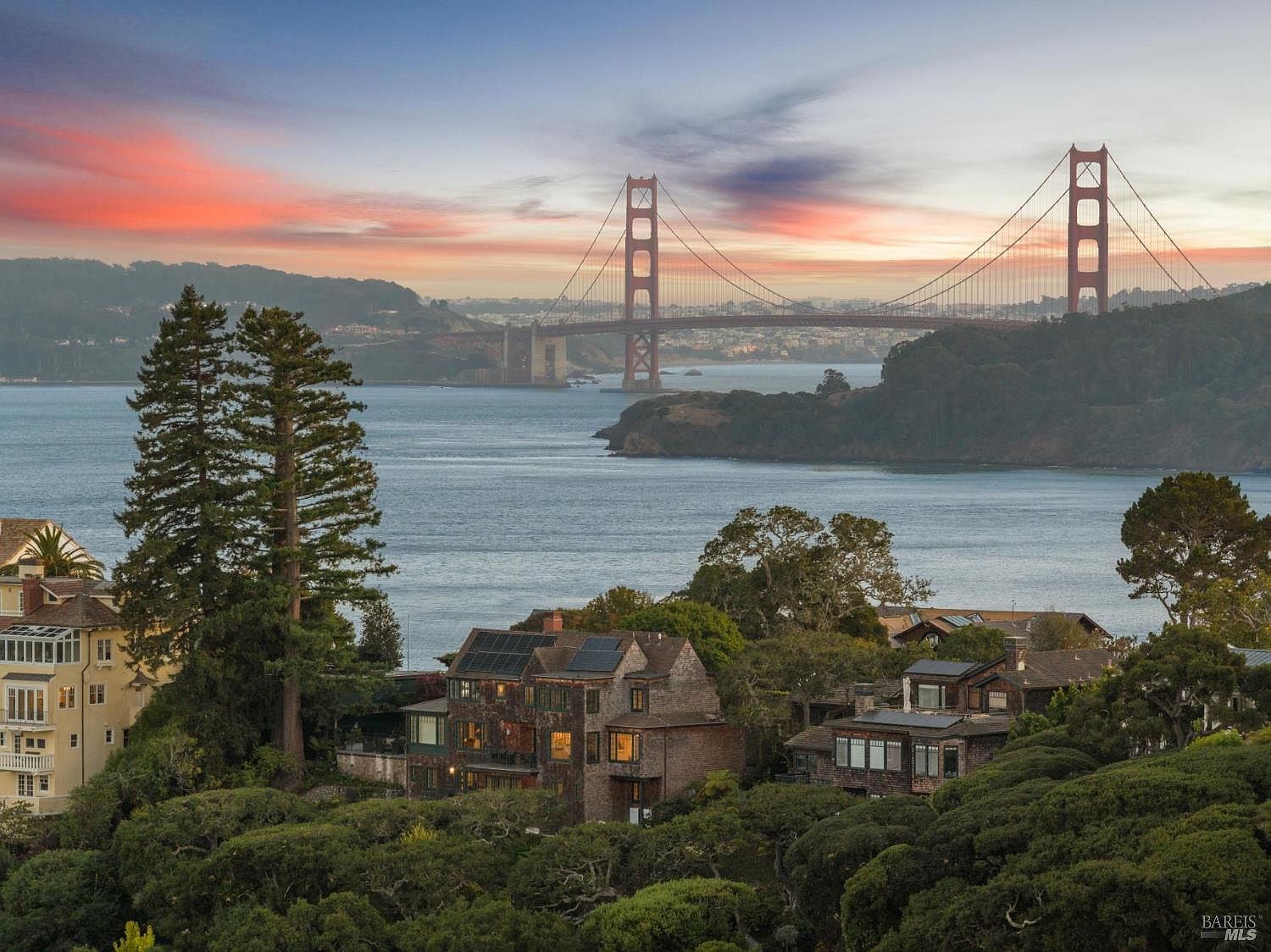 This image showcases a stunning front view of a multi-story home with a backdrop of the Golden Gate Bridge at sunset. The property features a mix of architectural styles, including wood siding and solar panels, nestled among lush greenery. The overall impression is one of luxury and tranquility, with a breathtaking view.
