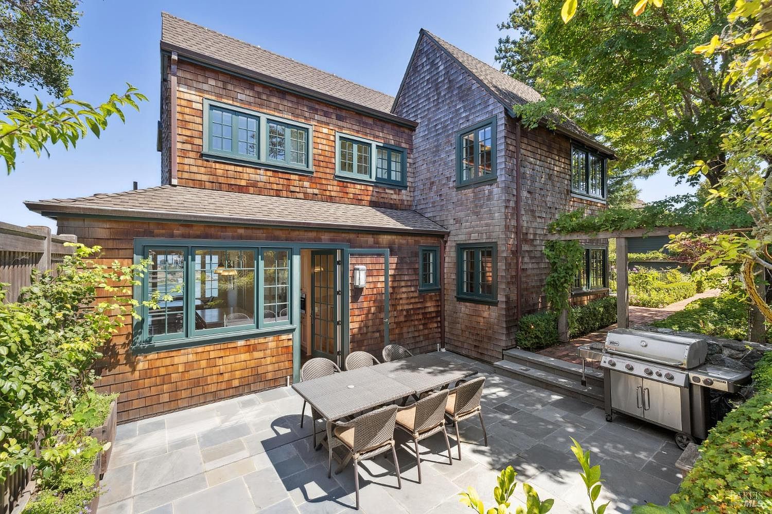 This image showcases a charming patio area adjacent to a cedar-shingled home. The patio features a stone tile surface, an outdoor dining set with seating for six, and a stainless steel grill. Lush greenery surrounds the space, creating a private and inviting outdoor living area.