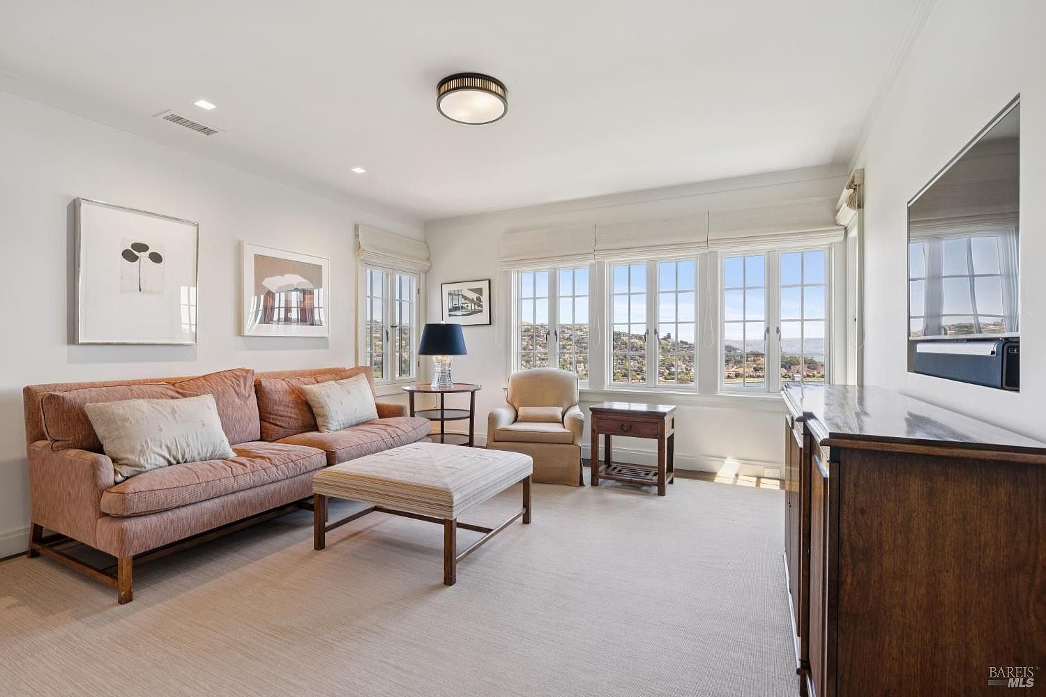 This is a bright and airy living room featuring a comfortable salmon-colored sofa with neutral pillows, a striped ottoman, and a beige armchair. Large windows offer a scenic view, complemented by artwork on the walls and a dark wood media console. The room is well-lit with a modern ceiling light fixture and natural light, creating a welcoming and sophisticated atmosphere.