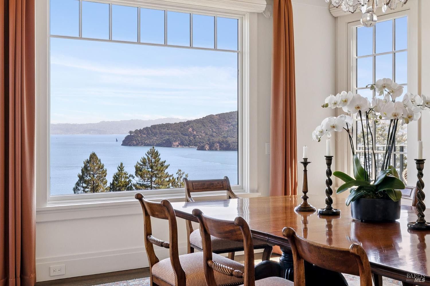 This is an interior shot of a dining room featuring a large wooden table with chairs. A large window offers a scenic view of the water and landscape, complemented by elegant curtains. The room is decorated with a chandelier and a floral arrangement, creating a sophisticated and inviting atmosphere.