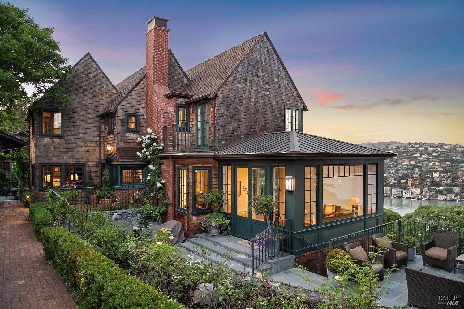 This is a rear exterior view of a charming multi-story house with a shingle exterior and a brick chimney. The house features a sunroom with large windows, a balcony, and a well-maintained garden with stone steps and lush greenery. The view overlooks a body of water and a hillside neighborhood, creating a picturesque setting.