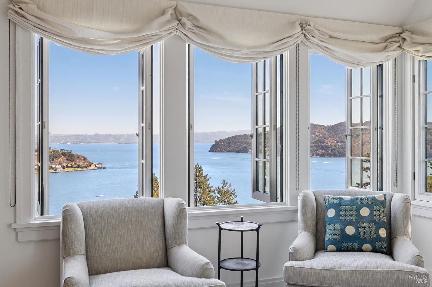 This interior shot showcases a cozy living room area with two armchairs positioned near a large window offering a scenic view of the water and distant hills. The room is bathed in natural light, highlighting the neutral color palette and creating a serene and inviting atmosphere. A small side table sits between the chairs, adding a touch of functionality.