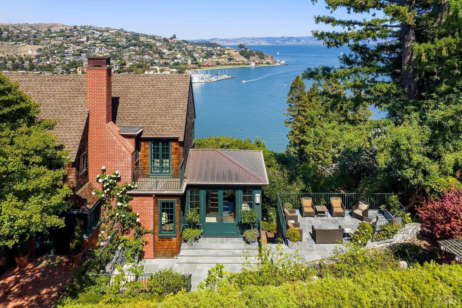 This aerial view showcases a charming home with a brick chimney and a well-maintained brown shingle roof. A cozy patio area with outdoor seating offers stunning views of the bay and surrounding landscape. The property is surrounded by lush greenery, providing a sense of privacy and tranquility.