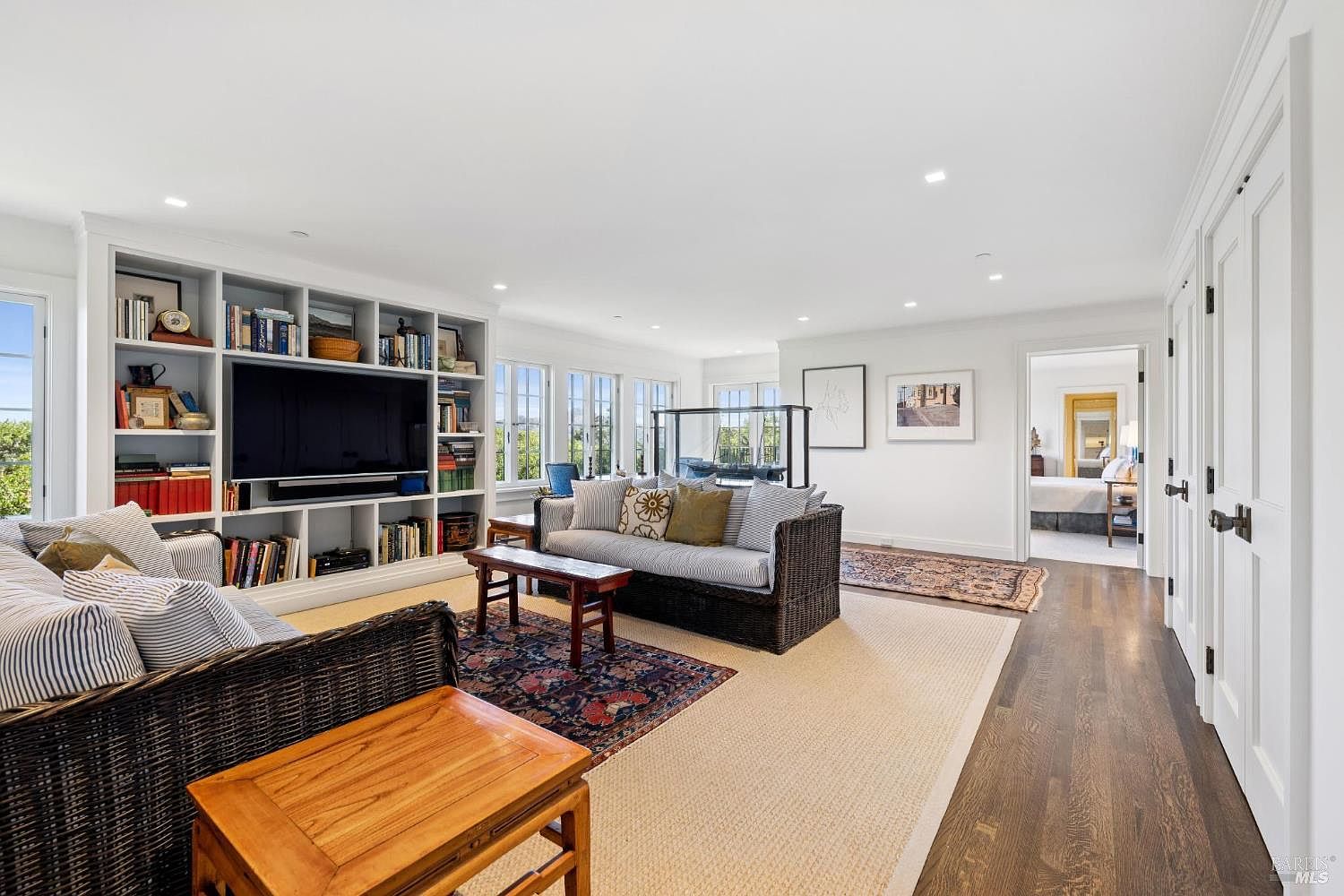 This is a spacious living room featuring a built-in bookshelf with a television, two wicker sofas, and wooden coffee tables. The room is well-lit with natural light from the windows and recessed lighting. The overall style is comfortable and inviting, perfect for relaxation and entertainment.