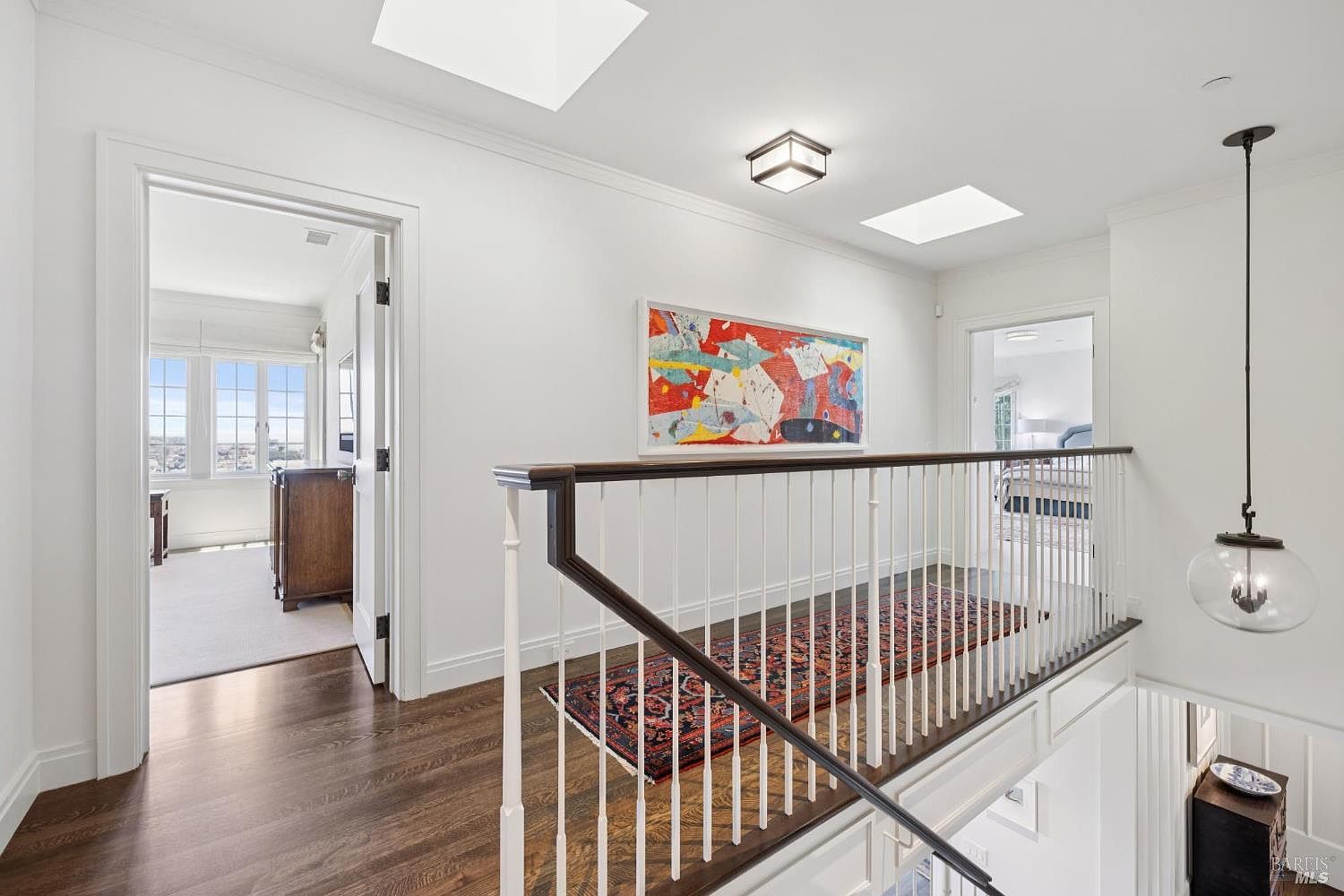 This is an interior shot of a hallway and staircase. The hallway features white walls, dark wood floors, and a colorful piece of artwork. A staircase with white railings and a dark wood banister leads to another level. A decorative rug is placed on the stairs. The space is well-lit with natural light from skylights and a hanging pendant light.