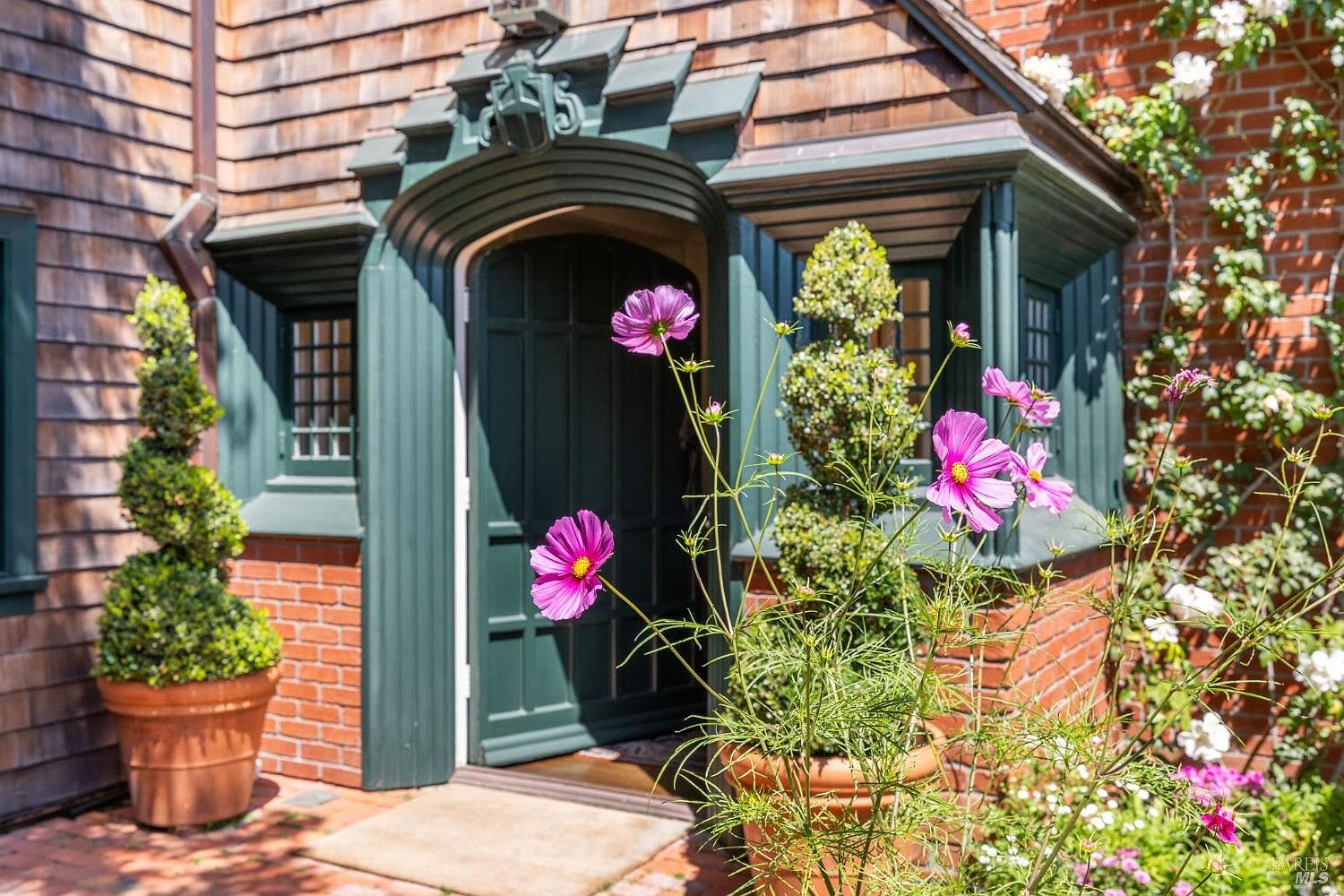 The image showcases an inviting entryway to a charming home. A dark green arched doorway is flanked by brick walls and lush greenery, including potted plants with pink flowers. The exterior features a mix of brick and wood shingles, creating a warm and welcoming aesthetic.