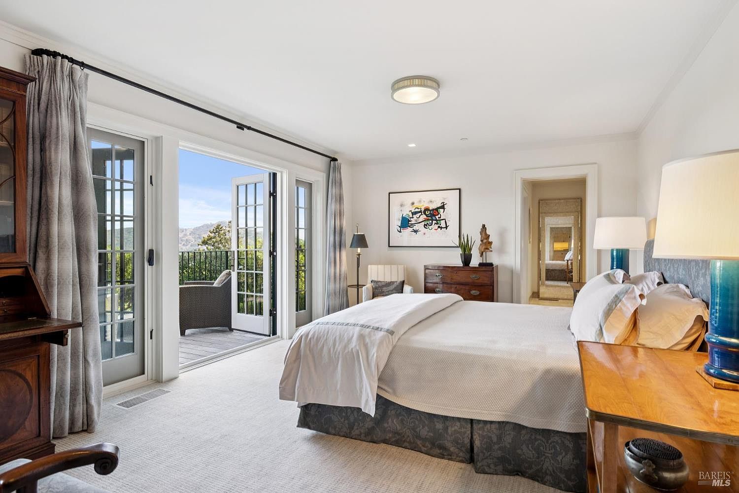 This is a bright and airy primary bedroom featuring a large bed with a white comforter and patterned bed skirt. French doors open to a balcony with a view of the surrounding landscape, bringing natural light into the room. The room is decorated with a mix of modern and traditional elements, including a dark wood desk and a contemporary art piece.
