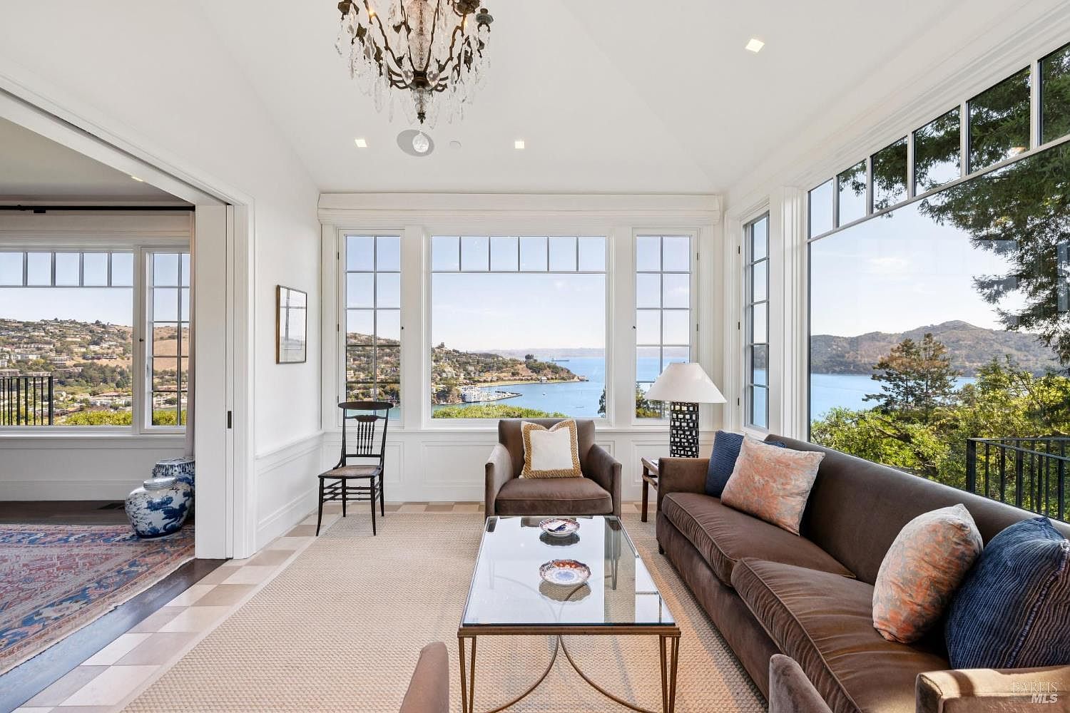 This is a bright and airy living room with large windows offering stunning water views. The room features a brown sofa with decorative pillows, a glass-topped coffee table, and a comfortable armchair. A chandelier hangs from the ceiling, adding a touch of elegance to the space.