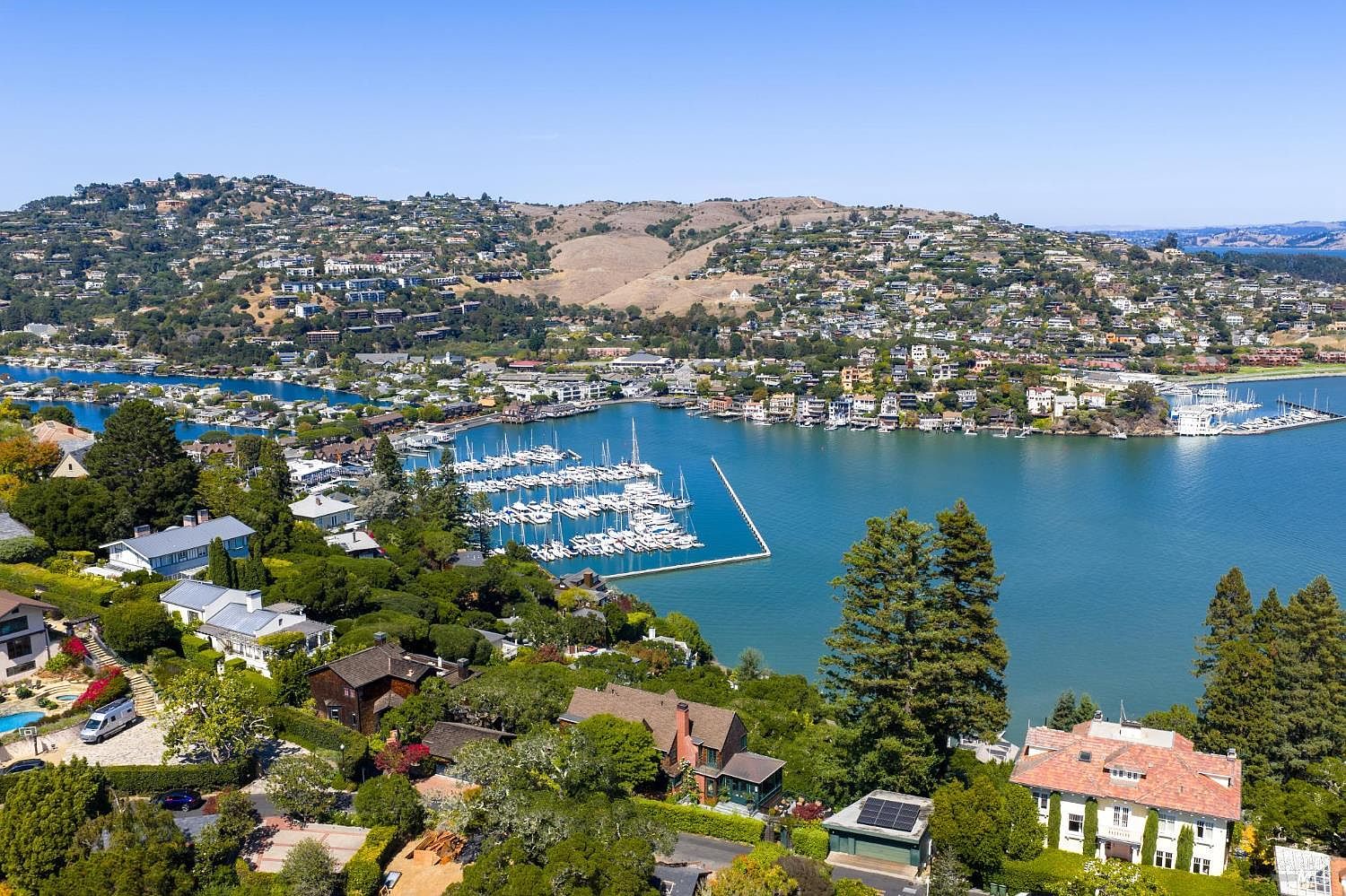 This aerial view showcases a stunning waterfront property nestled among lush greenery and overlooking a serene bay dotted with sailboats. The landscape features a mix of residential homes cascading down the hillside, creating a picturesque and desirable living environment. The overall impression is one of luxury, tranquility, and prime real estate.