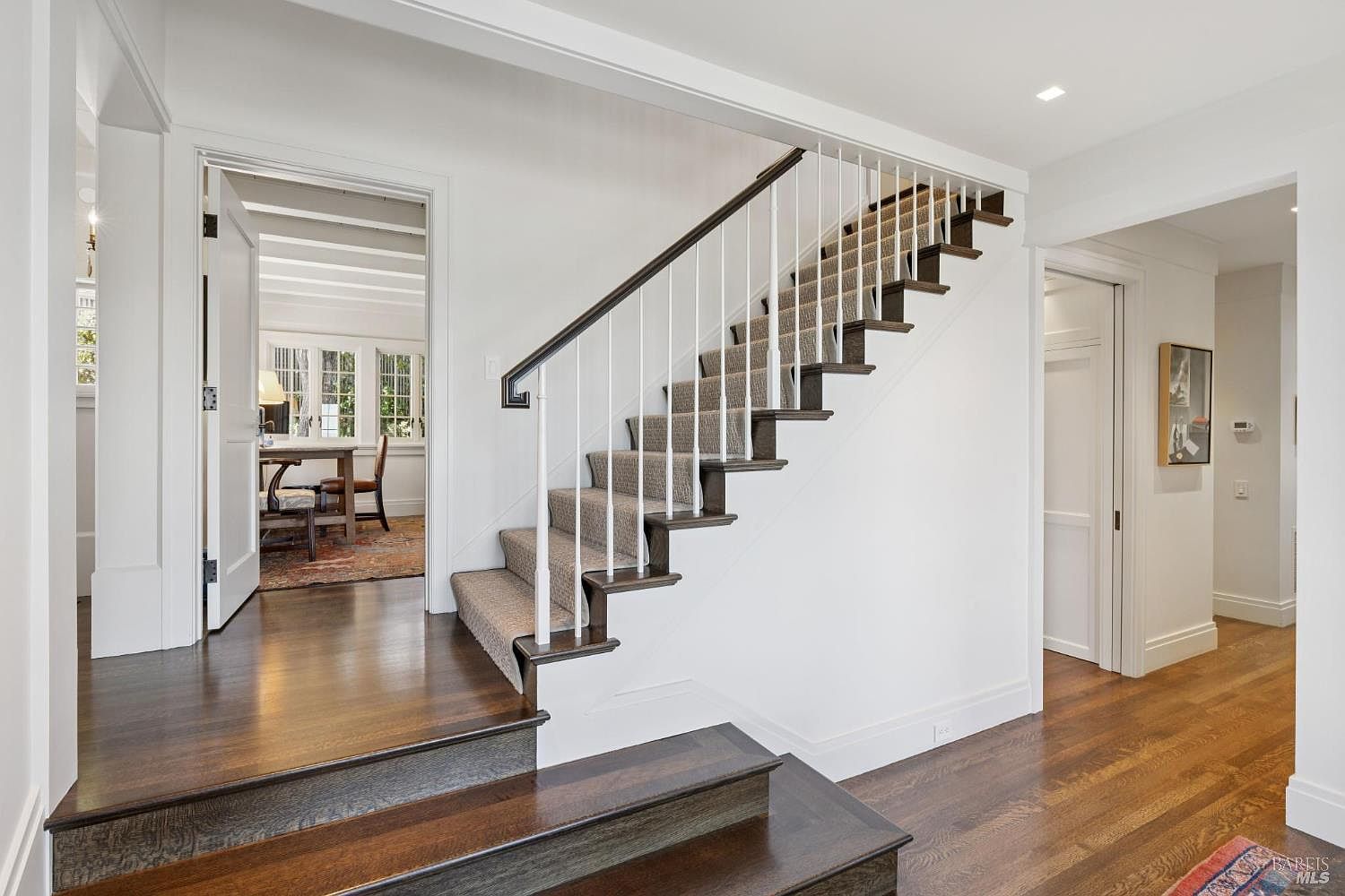 This interior shot showcases a well-lit hallway with a staircase. The staircase features dark wood steps with carpet and white railings, contrasting against the white walls. The hardwood flooring in the hallway adds warmth, and an open doorway reveals a glimpse into another room, enhancing the sense of space and flow.