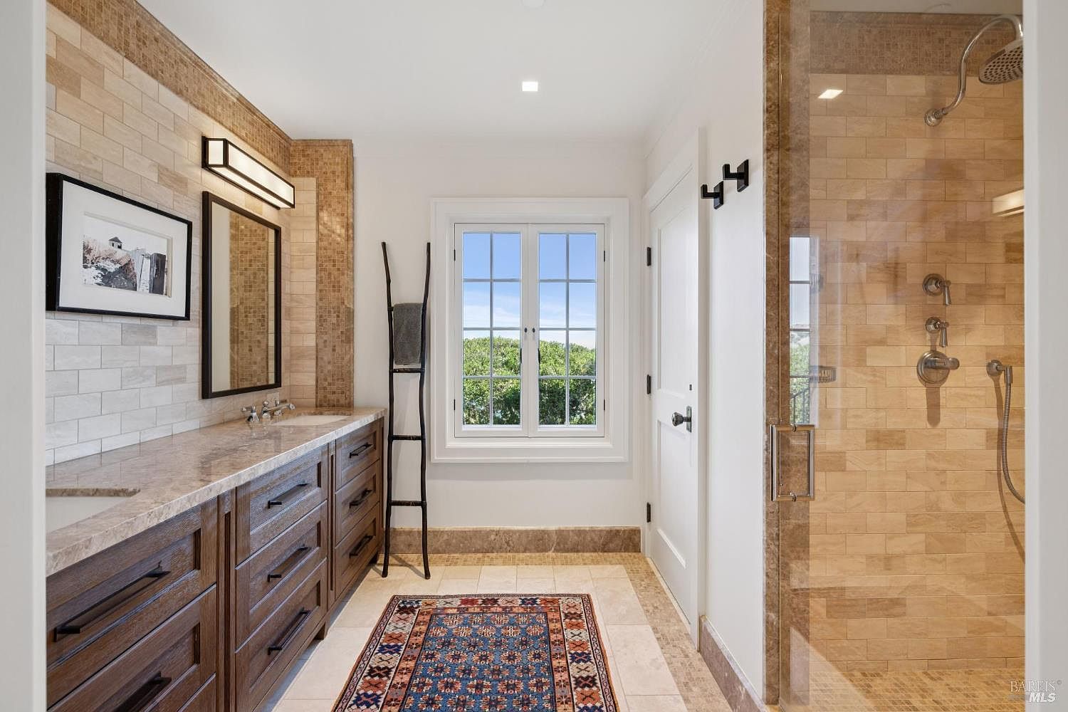 This is a well-lit primary bathroom featuring a long vanity with dark wood cabinetry and a light-colored countertop. A framed picture and a rectangular mirror hang above the vanity. A window provides natural light, and a decorative ladder leans against the wall. The shower is enclosed with glass and features beige tiling.