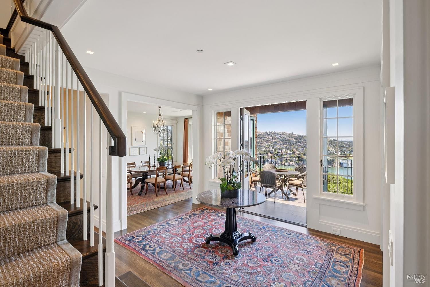 This interior shot showcases an elegant entryway featuring a staircase with a dark wood handrail and carpeted steps. A round table with a floral arrangement sits on a patterned rug, leading to an open dining room and a balcony with scenic views. The space is bright and airy, with white walls and trim, creating a welcoming and luxurious atmosphere.