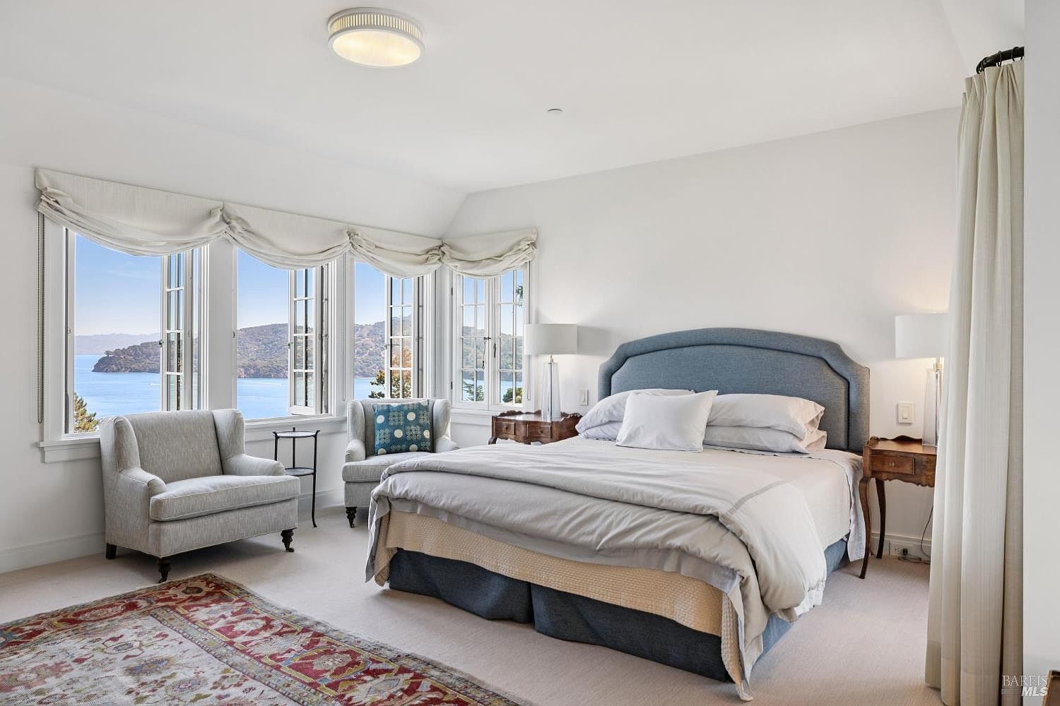 This is a bright and airy primary bedroom featuring a large bed with a blue headboard, neutral bedding, and two bedside tables with lamps. The room has large windows offering a scenic view of the water, and two armchairs are placed near the windows. The overall style is elegant and comfortable, creating a relaxing atmosphere.