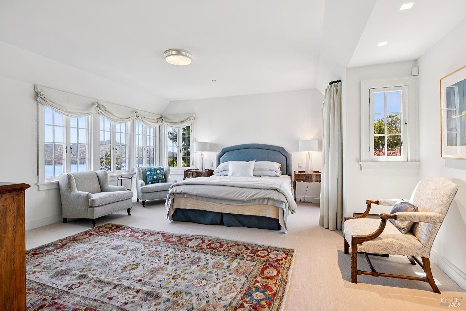 This is a bright and spacious primary bedroom featuring a large bed with a blue headboard, two armchairs near a bay window offering a scenic view, and an ornate rug. The room is painted in a clean white, enhancing the natural light, and includes bedside tables with lamps, creating a cozy and inviting atmosphere. A single chair sits near a window on the right.