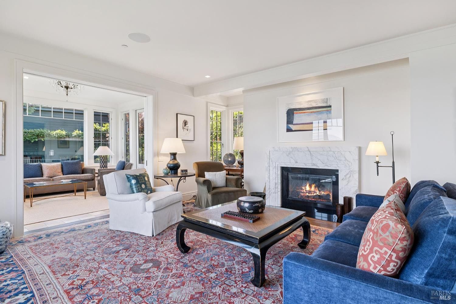 This is an interior shot of a living room featuring a fireplace, a blue velvet sofa with patterned pillows, and a patterned rug. The room has white walls and trim, with an open doorway leading to another seating area. The style is traditional with a touch of elegance, creating a warm and inviting atmosphere.