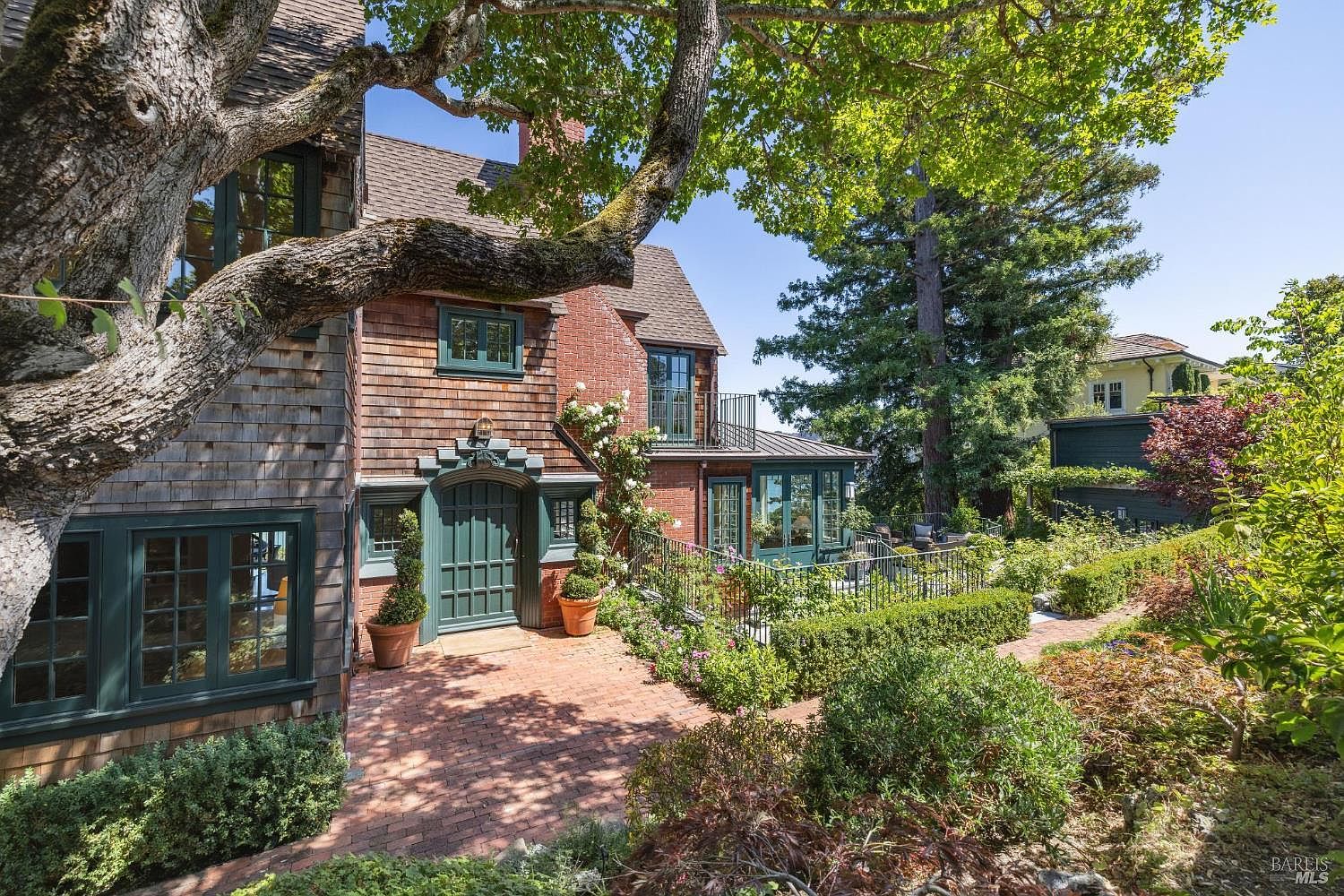 This image showcases the exterior of a charming home with a well-manicured yard and garden. The house features a mix of brick and shingle siding, complemented by dark green window frames and a matching front door. A brick pathway leads to the entrance, surrounded by lush greenery and flowering plants, creating an inviting and picturesque setting.