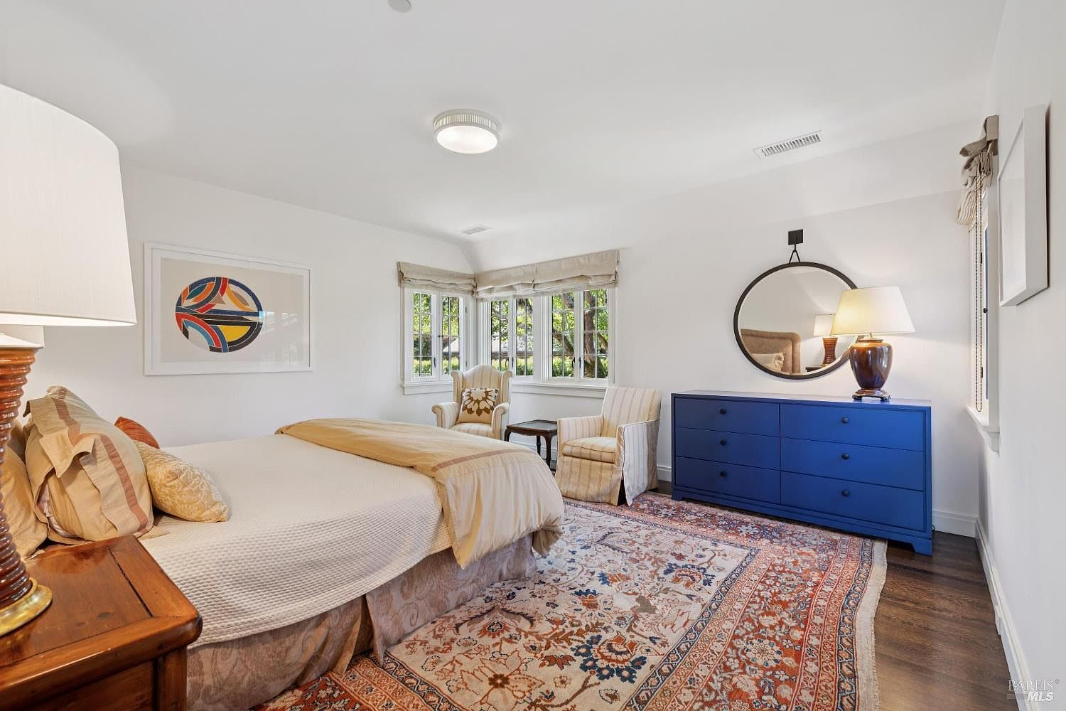 This is a well-lit primary bedroom featuring a bed with neutral-toned bedding, a blue dresser with a round mirror above it, and two armchairs near a window. The room is decorated with a colorful rug and artwork, creating a comfortable and inviting atmosphere. The perspective is from the foot of the bed, showcasing the room's layout and design elements.