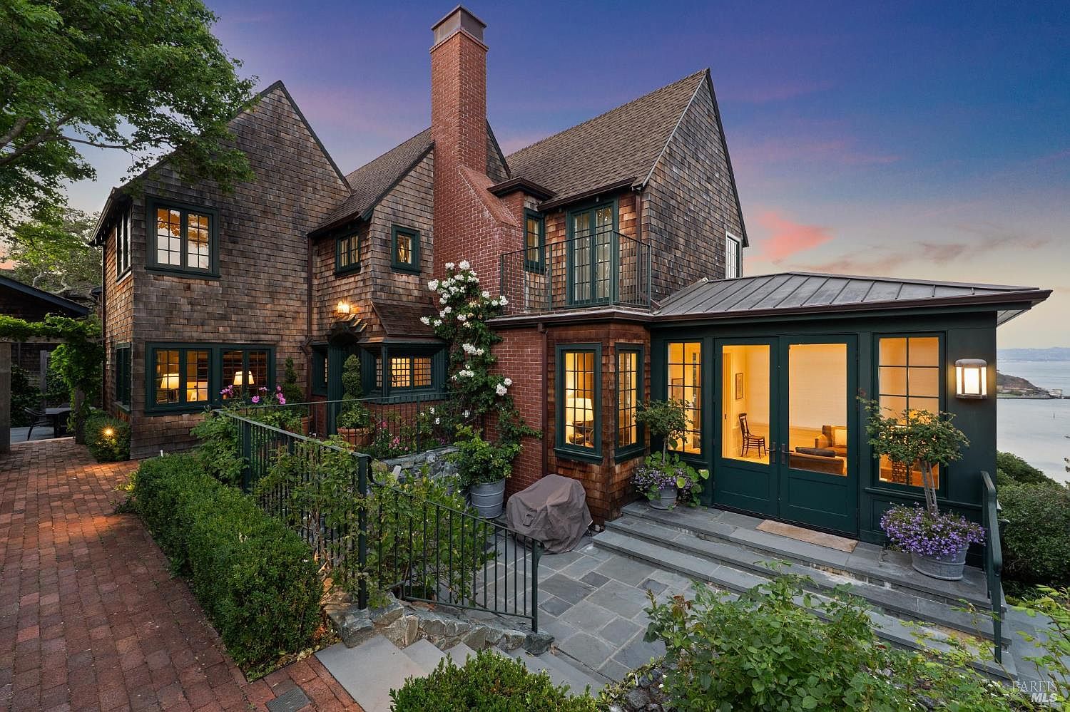 This is a rear exterior view of a multi-story home with a shingle exterior and dark green trim. The property features a brick chimney, a small balcony, and a sunroom with large windows. The landscaping includes a stone patio, garden beds, and mature trees, creating a serene and private outdoor space.