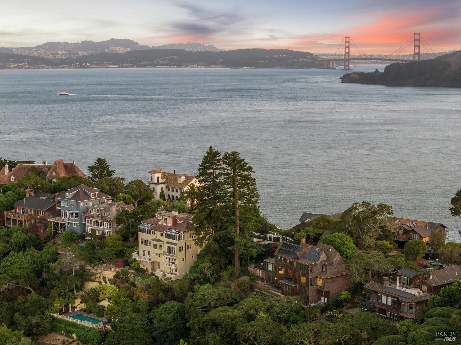 This aerial view showcases a collection of luxury homes nestled among lush greenery, overlooking a serene bay with the Golden Gate Bridge in the distance. The properties feature diverse architectural styles, including traditional and modern designs, with well-maintained landscaping and possible amenities like pools. The overall impression is one of exclusivity and stunning natural beauty.
