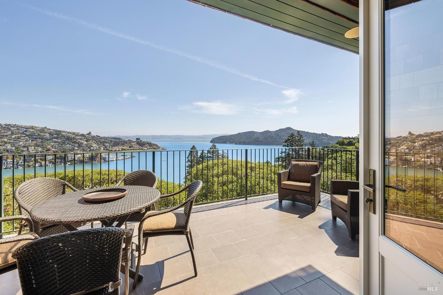 This image showcases a spacious balcony with outdoor furniture, including a round table with chairs and two armchairs. The balcony features a black metal railing and a tiled floor. The view from the balcony overlooks a scenic body of water with hills and trees in the background, creating a serene and inviting outdoor living space.