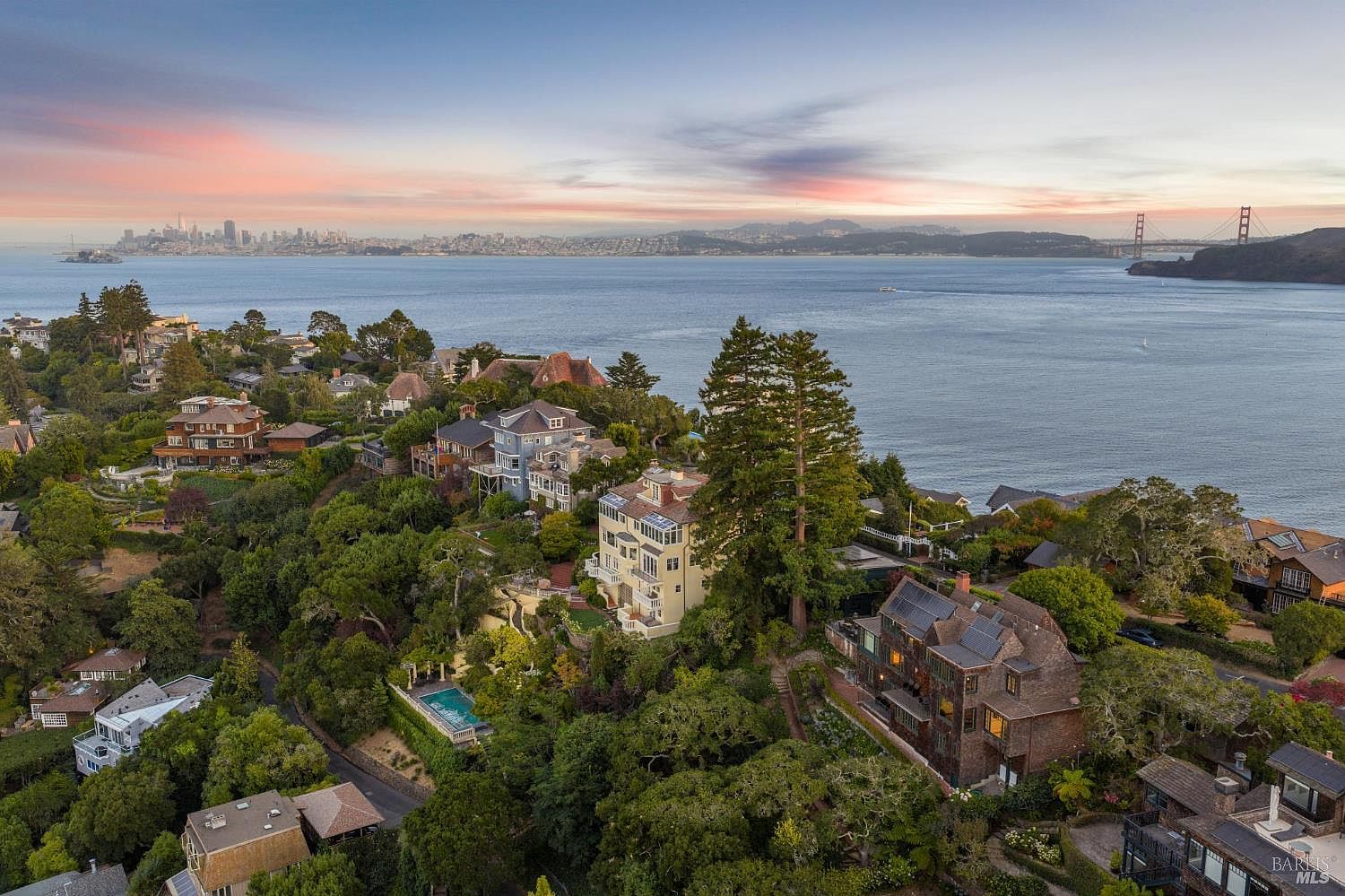 This aerial view showcases a collection of luxury homes nestled among lush greenery, overlooking a serene bay with a cityscape and the Golden Gate Bridge in the distance. The properties feature diverse architectural styles, some with pools and well-manicured gardens, creating an impression of exclusivity and tranquility. The scene is bathed in the soft light of either sunrise or sunset, enhancing the picturesque setting.
