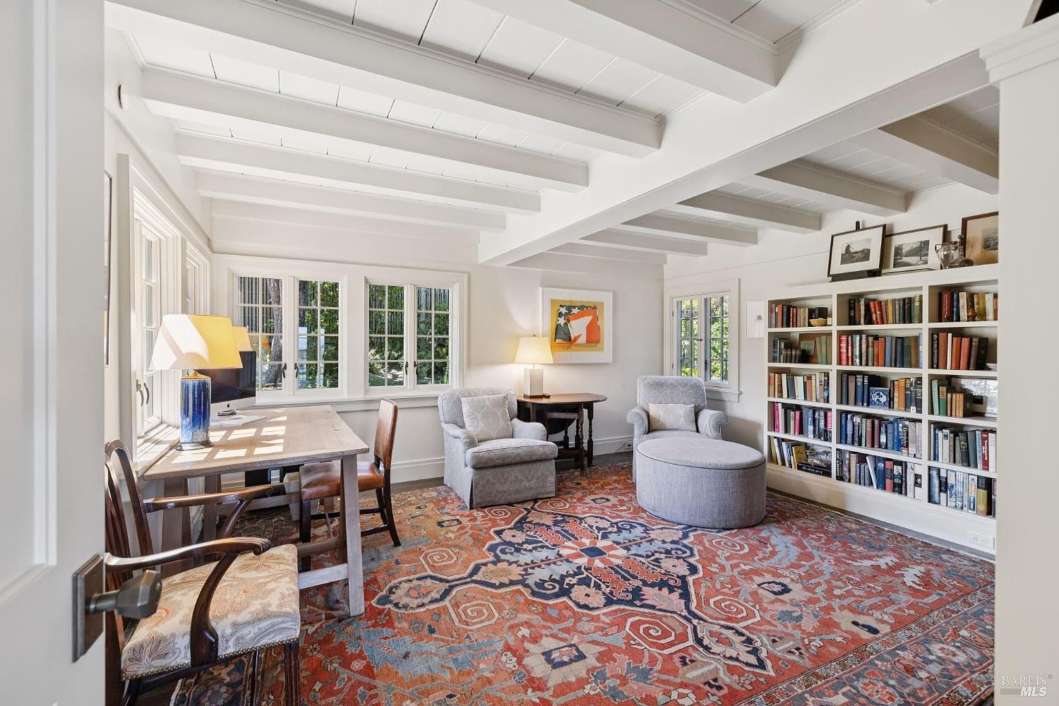 This is an interior shot of a well-lit office or study. The room features a large area rug, a desk with a computer, comfortable seating, and a built-in bookcase filled with books. The ceiling has exposed beams, and there are multiple windows providing natural light, creating a cozy and inviting workspace.