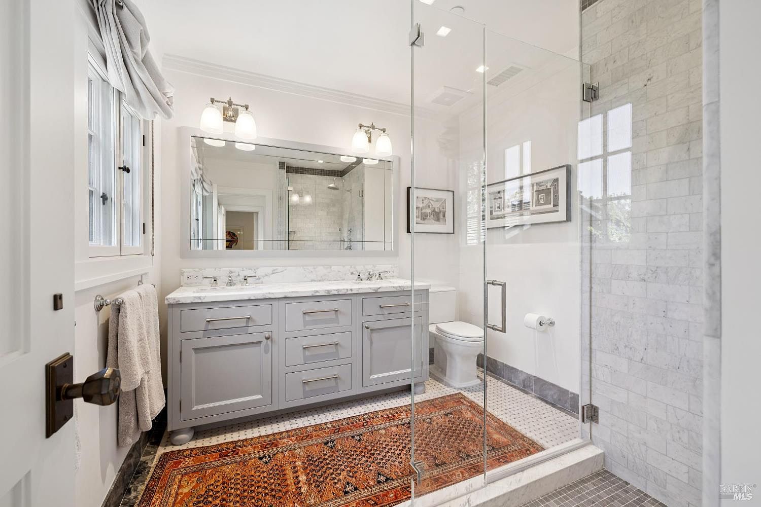 This is a well-lit primary bathroom featuring a double vanity with a marble countertop and gray cabinetry. A large mirror hangs above the vanity, reflecting the shower area. The bathroom includes a glass-enclosed shower and a toilet, with an orange patterned rug adding a touch of warmth to the tiled floor.