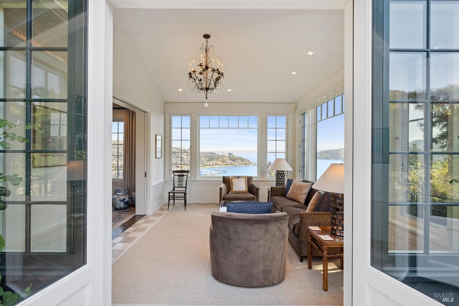 This is an interior shot of a sunroom or living room featuring a stunning water view through large windows. The room is elegantly furnished with a brown sofa, armchairs, and a chandelier, creating a sophisticated and inviting atmosphere. The open doors on either side of the room provide a sense of spaciousness and connection to the outdoors.