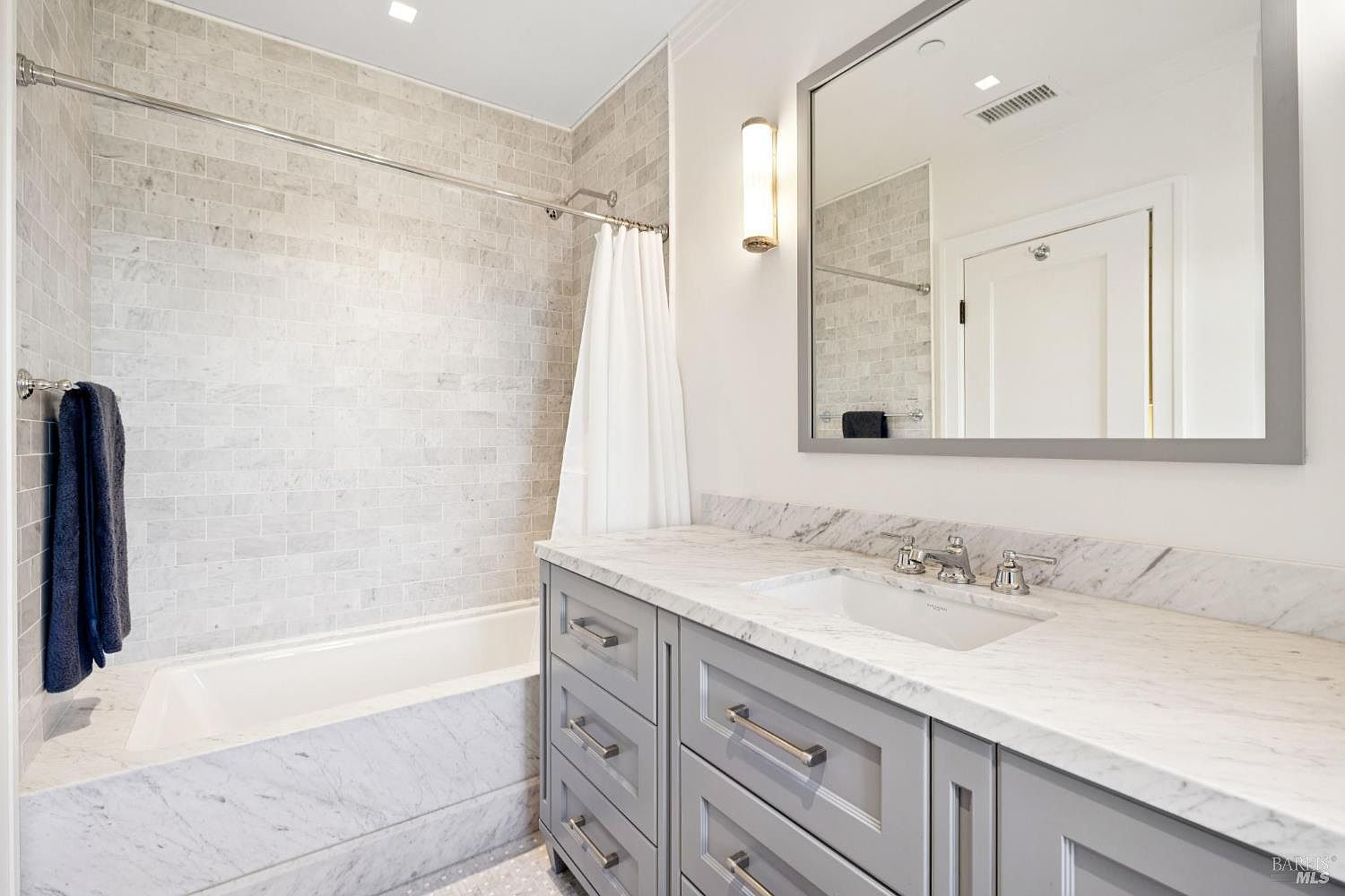 This is a well-lit bathroom featuring a bathtub with light gray marble-like tiling, a white shower curtain, and a gray vanity with a white marble countertop. A large mirror hangs above the sink, reflecting the bathroom's clean and modern design. The overall impression is one of understated elegance and functionality.