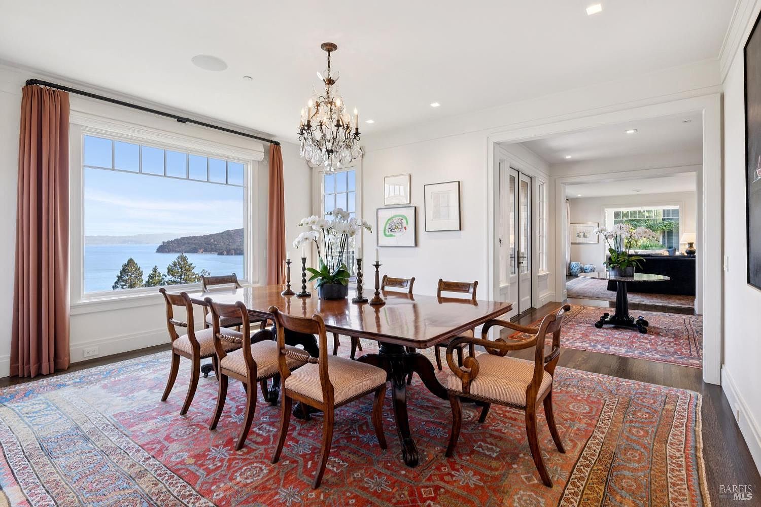 This is an interior shot of a dining room featuring a large wooden dining table with chairs, a crystal chandelier, and a large window with a view of the water. The room is decorated with a traditional rug and artwork, creating an elegant and inviting atmosphere. The perspective is from the corner of the room, capturing the table setting and the view.