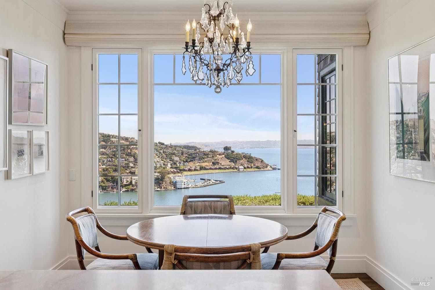 This is an interior shot of a dining room featuring a round wooden table surrounded by four chairs with woven backs and light blue cushions. A crystal chandelier hangs above the table, and a large window provides a scenic view of the water and hillside homes. The room is bright and airy, with white walls and trim, creating an elegant and inviting atmosphere.