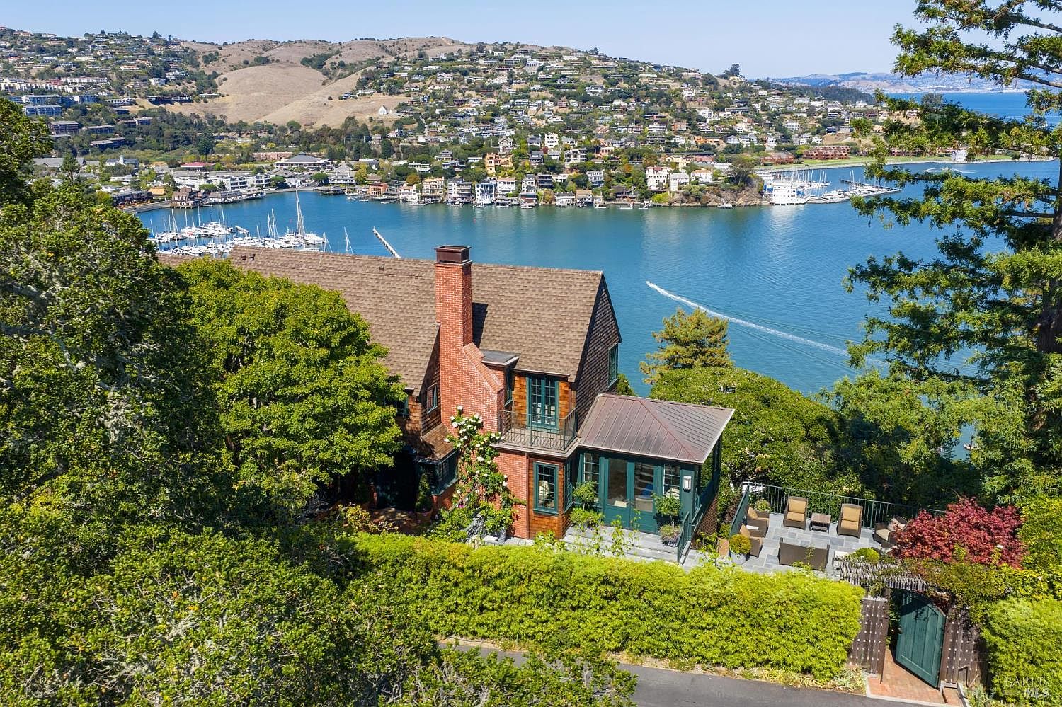 This aerial view showcases a charming home with a brick chimney and a brown roof, nestled amidst lush greenery overlooking a serene bay. A patio area with outdoor seating adds to the appeal, while the surrounding landscape features a mix of trees, hedges, and a glimpse of the road, creating a picturesque and private setting. The overall impression is one of tranquility and natural beauty.