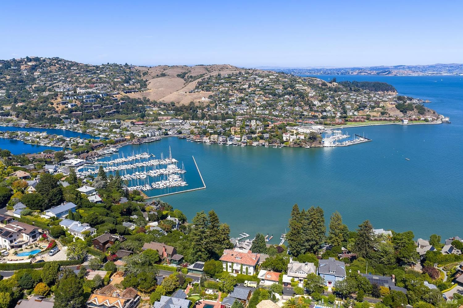 This aerial shot showcases a picturesque coastal community with a marina filled with sailboats. The landscape features hillside homes nestled among lush greenery, overlooking a serene bay. The overall impression is one of luxury and tranquility, highlighting the desirable waterfront location.