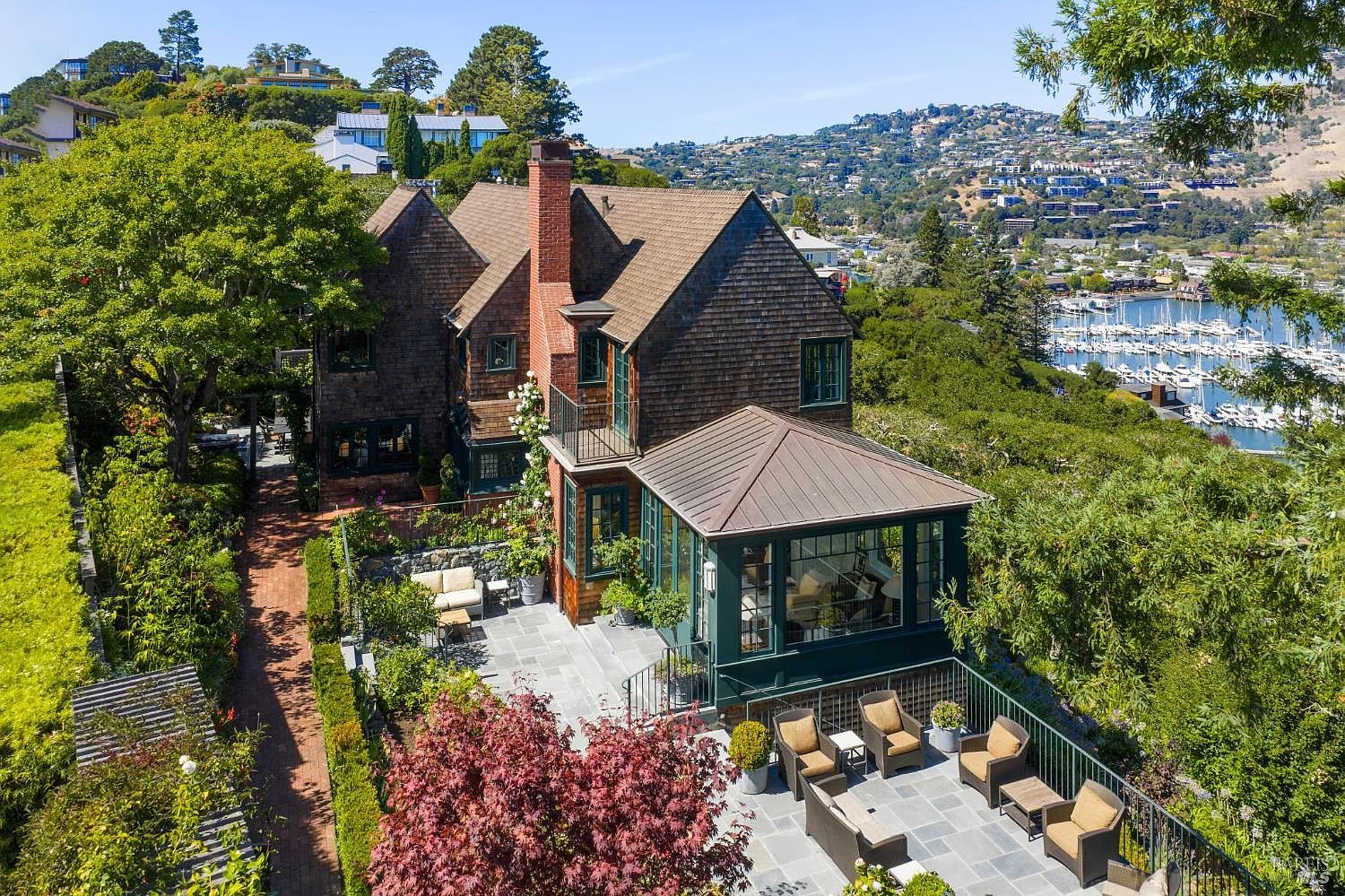 This aerial view showcases a charming multi-story home with a shingle exterior and a brick chimney. The property features a well-manicured garden with a patio area furnished with outdoor seating. In the background, a scenic view of a harbor filled with boats and rolling hills adds to the property's appeal.