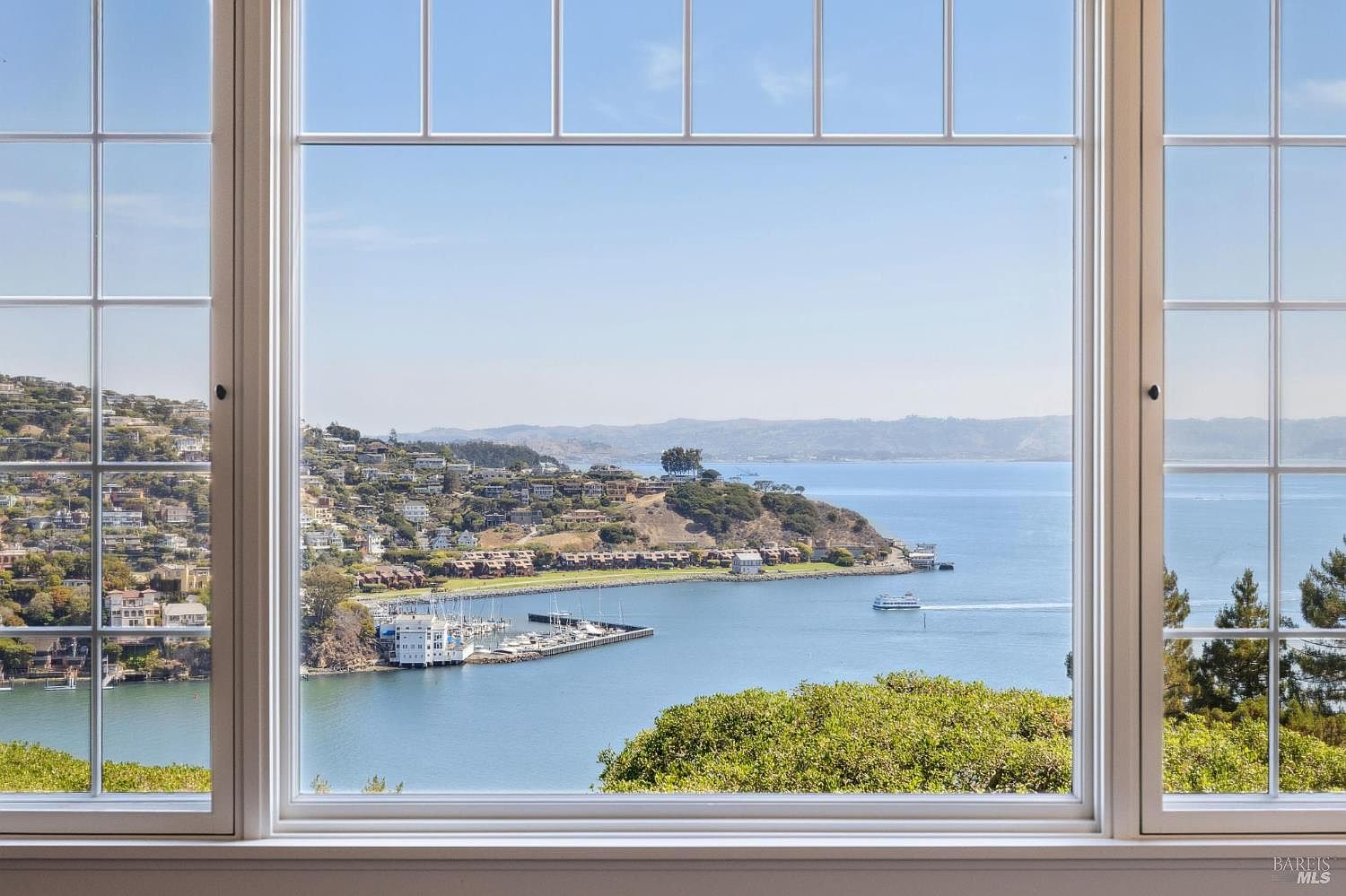 This image showcases a stunning view of the bay through a large, multi-paned window, likely from a living room. The window frames the picturesque scenery, featuring a coastline dotted with houses, a marina, and boats sailing on the water. The overall impression is one of tranquility and luxury, emphasizing the property's desirable location and breathtaking views.
