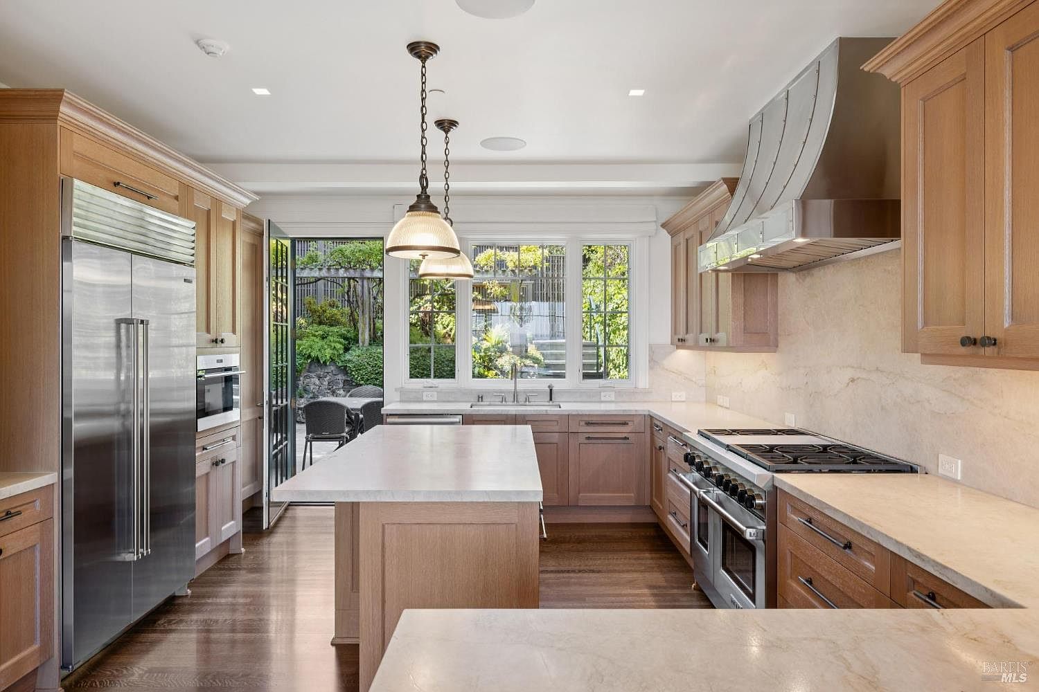 This is a well-lit kitchen featuring light wood cabinetry, stainless steel appliances, and light-colored countertops. A large island provides ample workspace, and pendant lights hang above it. The kitchen opens to an outdoor area through glass doors, creating a seamless indoor-outdoor flow.