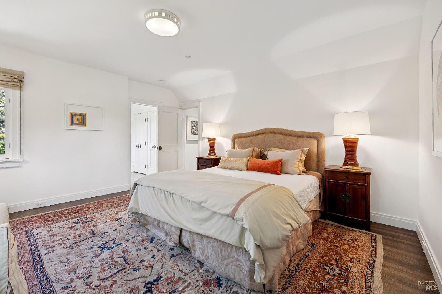 This is a well-lit primary bedroom featuring a large bed with a patterned headboard and neutral-toned bedding. Two dark wood nightstands with lamps flank the bed, and a colorful area rug covers most of the hardwood floor. The walls are painted white, and there is a doorway leading to another room, creating a bright and inviting space.