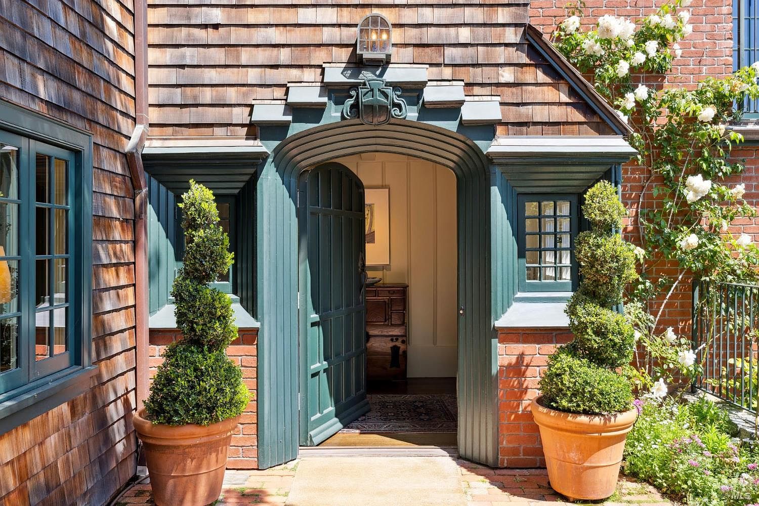 The image showcases an inviting entryway to a home, featuring a unique arched doorway with teal-painted wood and brick accents. Two potted spiral topiaries flank the entrance, adding a touch of greenery and elegance. The exterior combines wood shingles and brick, creating a charming and welcoming aesthetic.