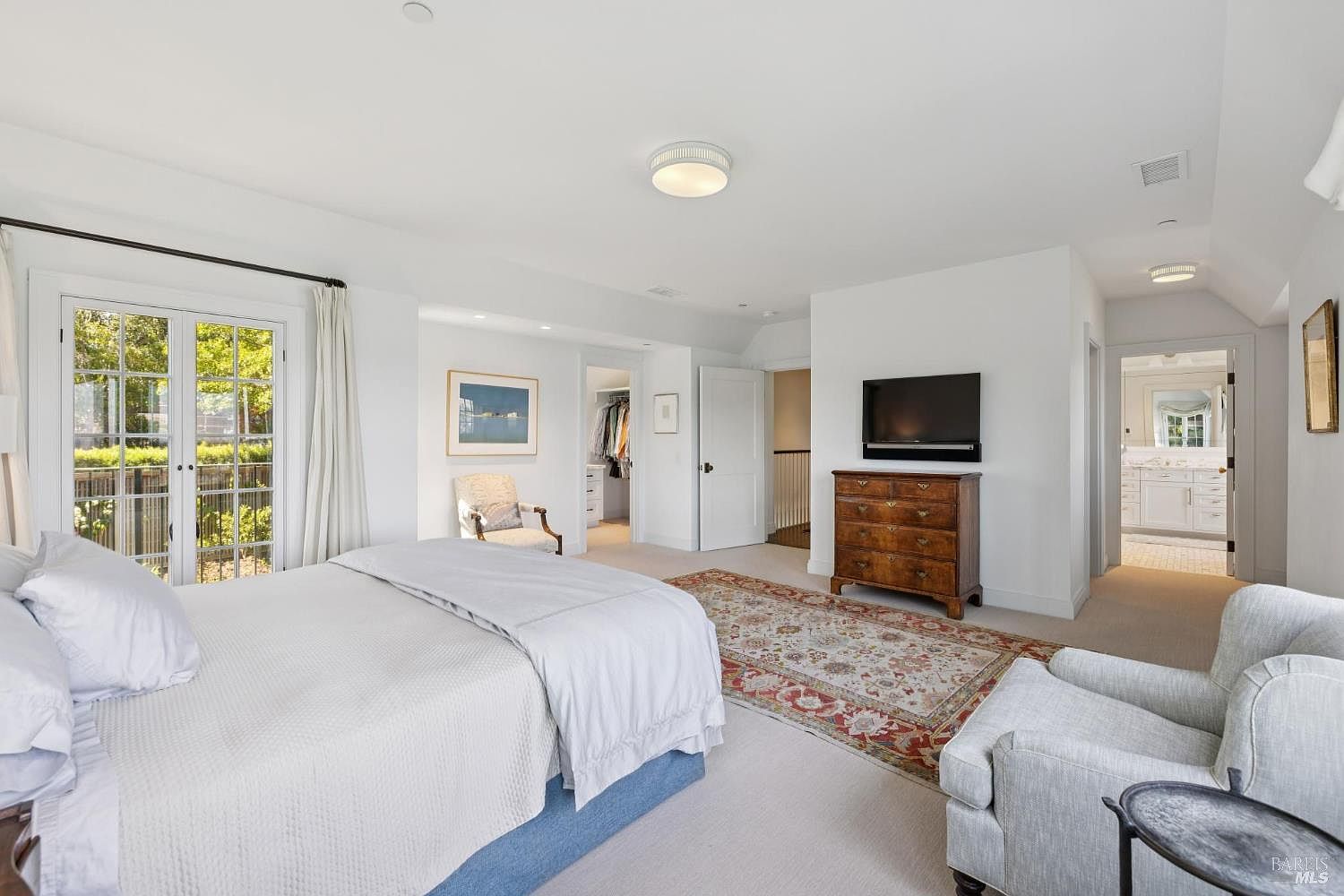 This is a bright and spacious primary bedroom featuring a large bed with white linens, a wooden dresser with a wall-mounted TV above it, and a cozy armchair. The room has white walls, a neutral carpet, and a decorative rug, creating a serene and inviting atmosphere. French doors lead to an outdoor area, enhancing the room's appeal.