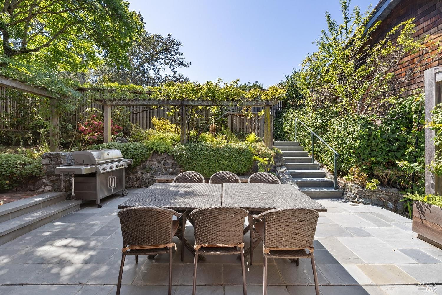 This image showcases a beautifully landscaped backyard featuring a stone patio with a large outdoor dining table and chairs. A stainless steel grill sits to the side, and a pergola covered in vines adds a touch of elegance. Stone steps lead up to a higher level of the garden, creating a multi-tiered outdoor space perfect for entertaining and relaxation.